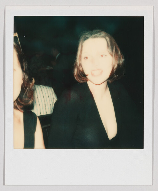 Young woman in a low-cut black dress smiles toward the camera at a dimly lit event.