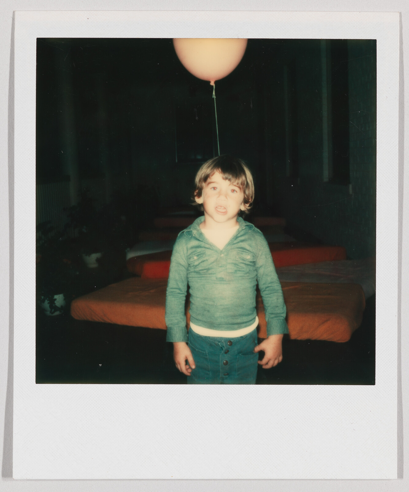 Young child standing indoors with a pink balloon tied to their head, beds with orange covers in background.