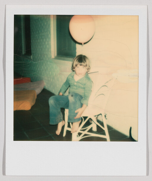 A young child sits barefoot on a white chair indoors with a balloon floating above their head.