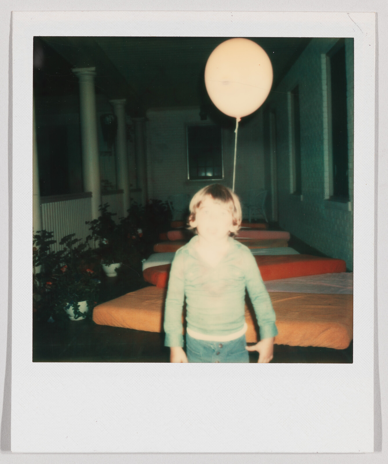 A young child stands indoors holding a balloon, with several orange-covered beds in the background.