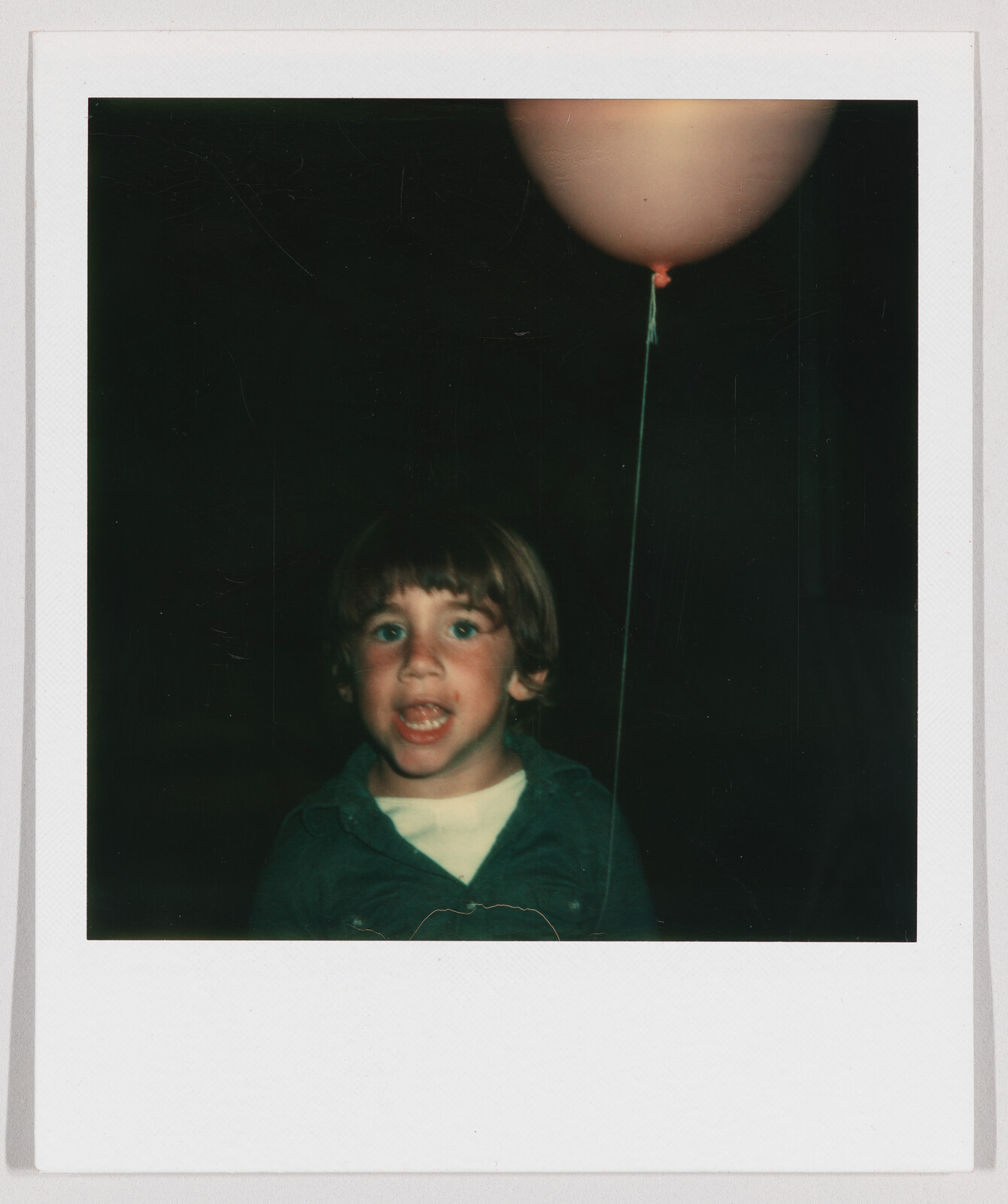 Young child with an open mouth holding a pink balloon on a string against a dark background.