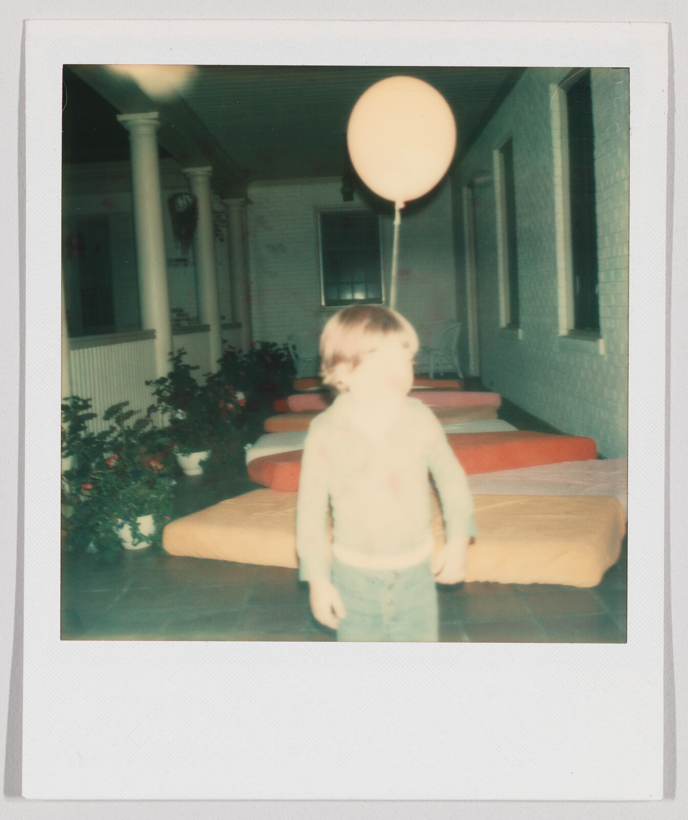 A young child stands holding a balloon, with several mattresses lined up behind them on a porch.