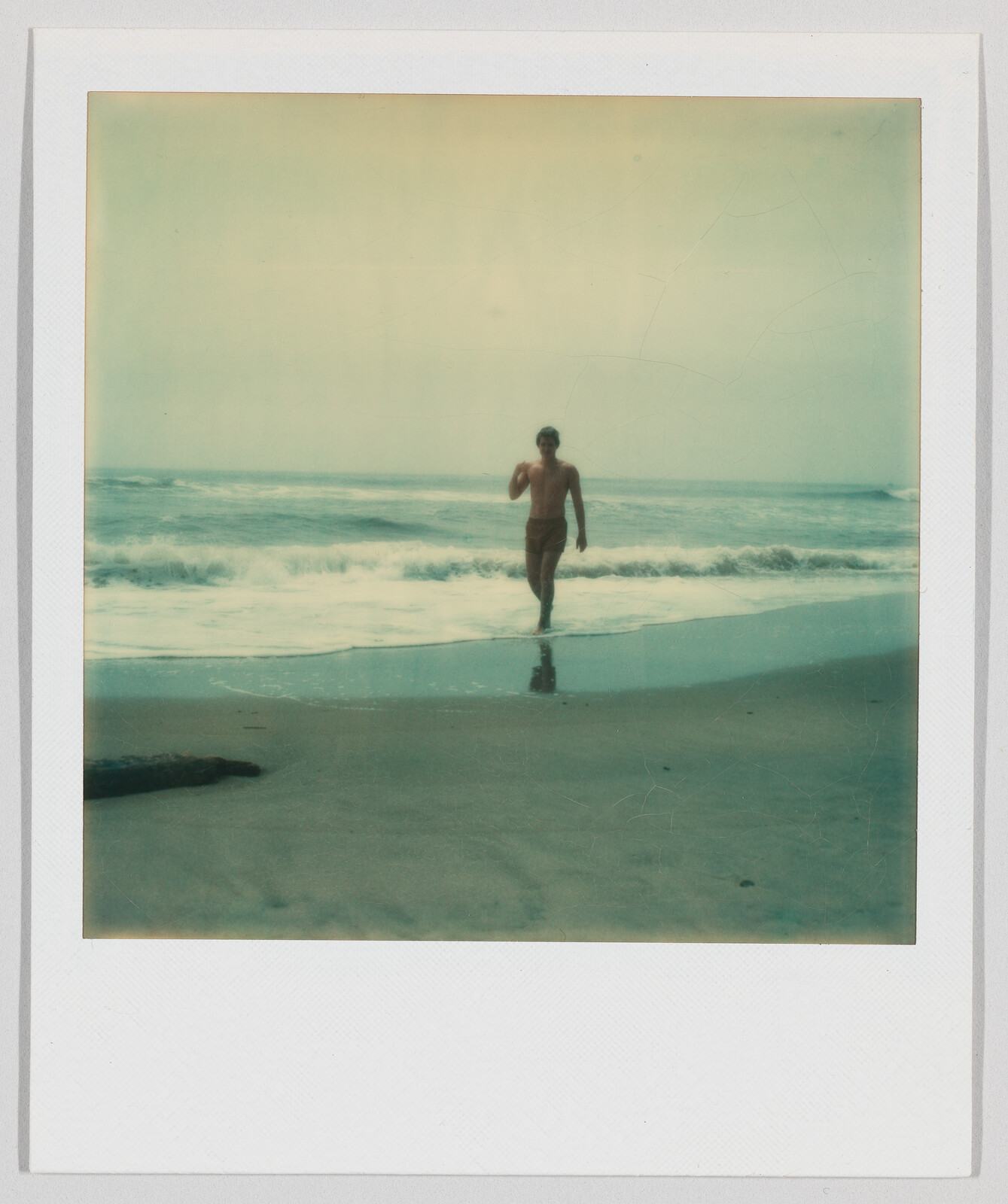A person in swim trunks walks out of the ocean onto a sandy beach with gentle waves.