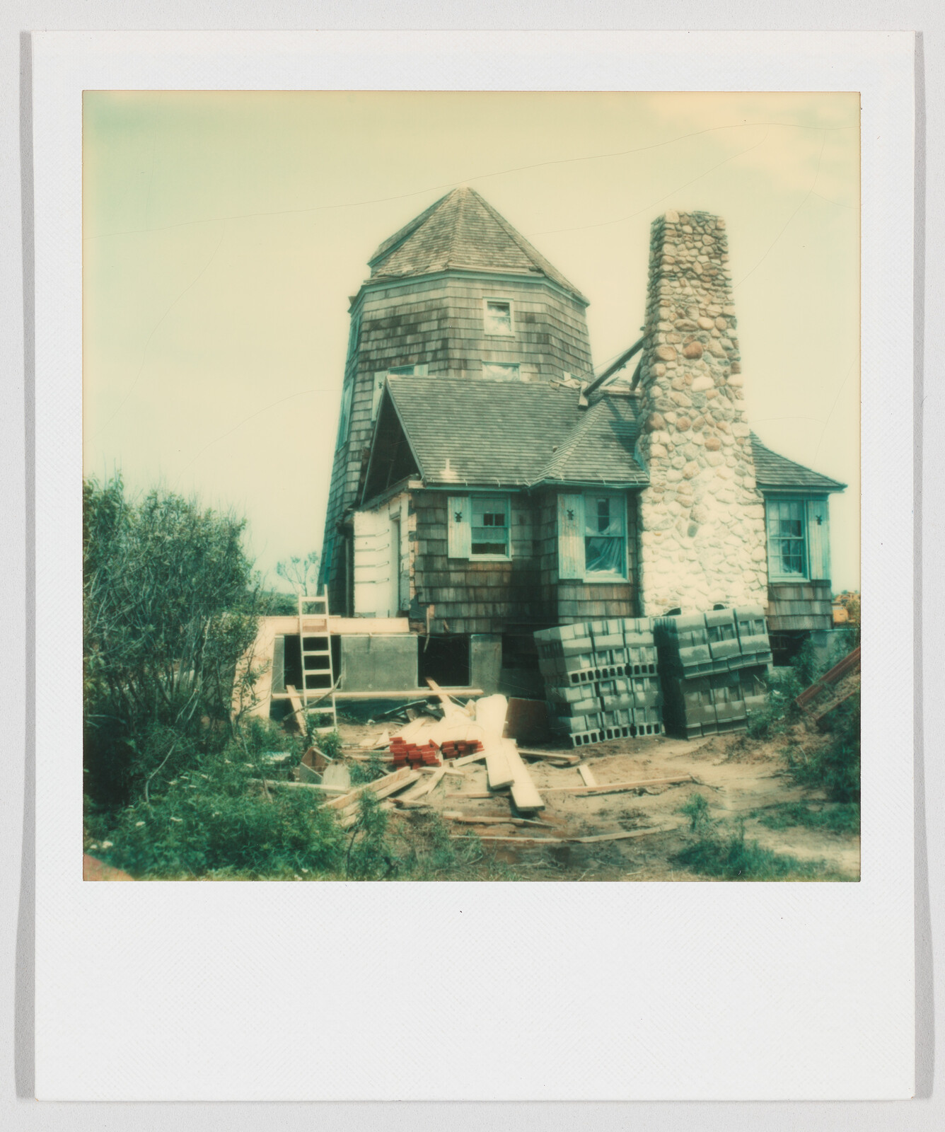 A small wooden house with a stone chimney, surrounded by construction materials and stacks of cinder blocks.