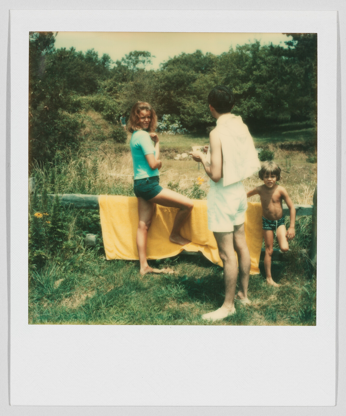 A woman, a man, and a young boy stand outside near a yellow towel hanging on a wooden fence.