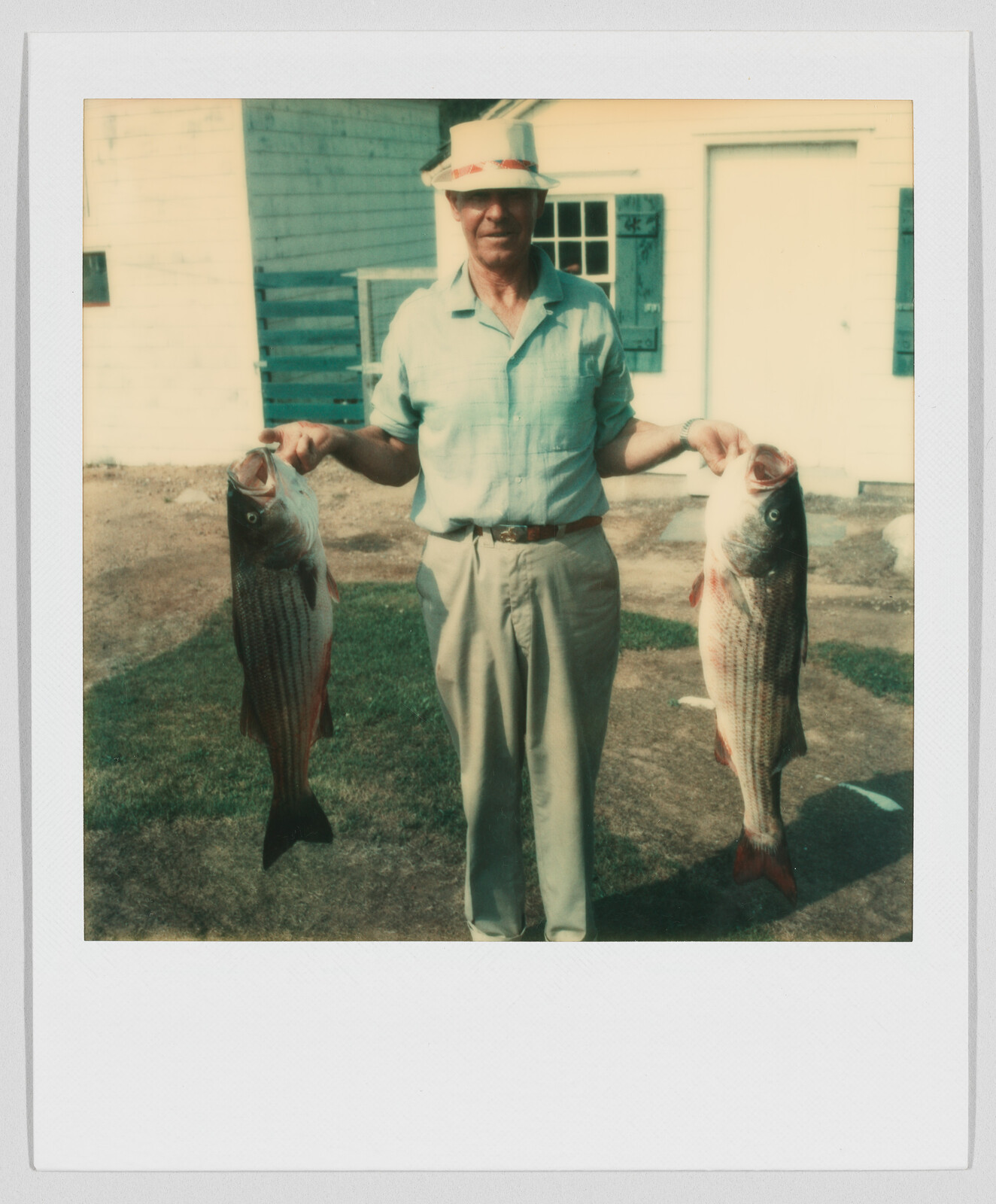 Man standing outside holding two large fish, one in each hand, with a house in the background.