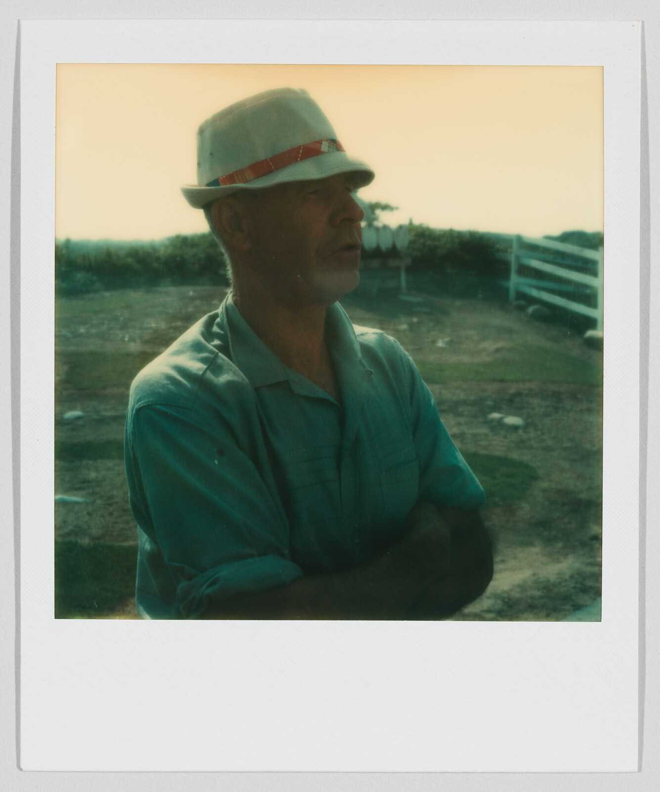 Man wearing a light-colored hat and shirt stands outdoors with arms crossed, looking to the side.