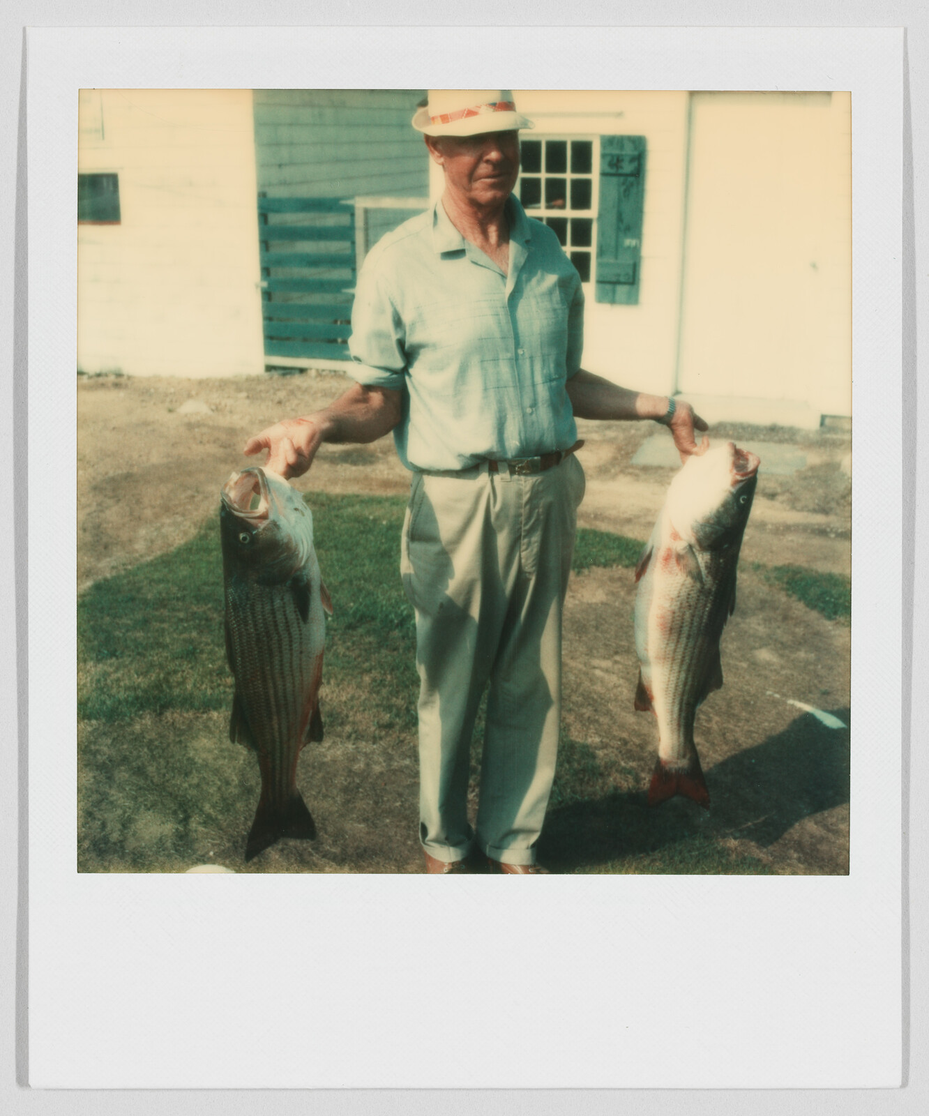 Man wearing a hat stands outside holding two large fish, one in each hand, by their tails.