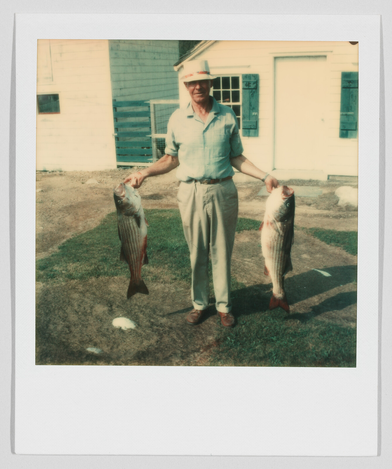 Older man standing outside, smiling and holding two large fish, one in each hand, near a house.