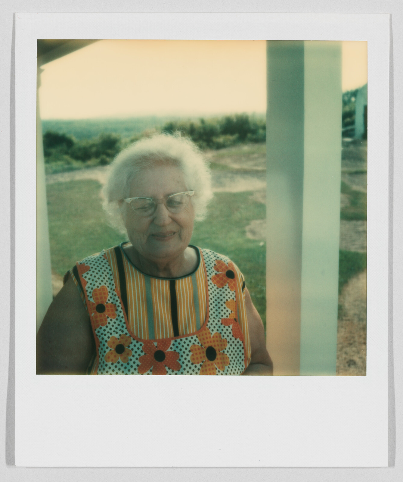 Elderly woman with white hair and glasses stands outside, wearing a colorful dress with orange flowers.