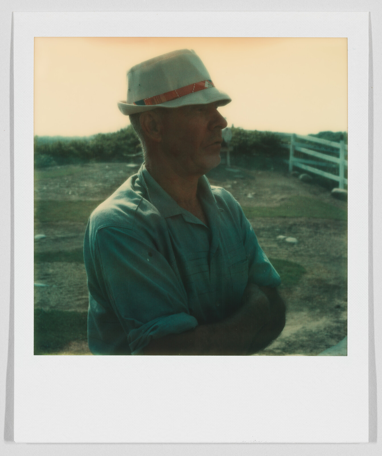Man wearing a light-colored hat and shirt stands outdoors with arms crossed, looking to the side.