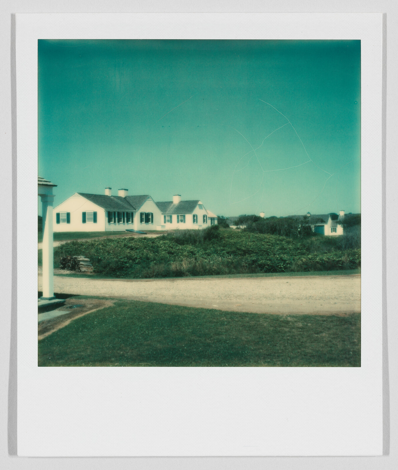 Several white houses with dark roofs sit behind a grassy area and a dirt road under a clear blue sky.