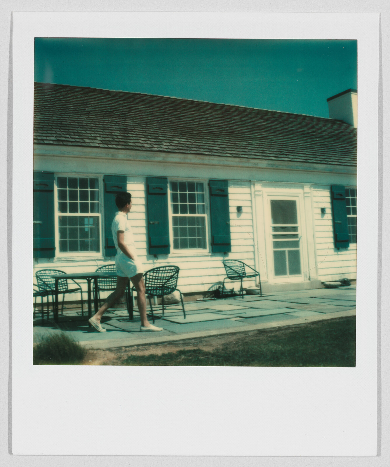 Person in white clothes walking past outdoor chairs and tables in front of a white house with blue shutters.