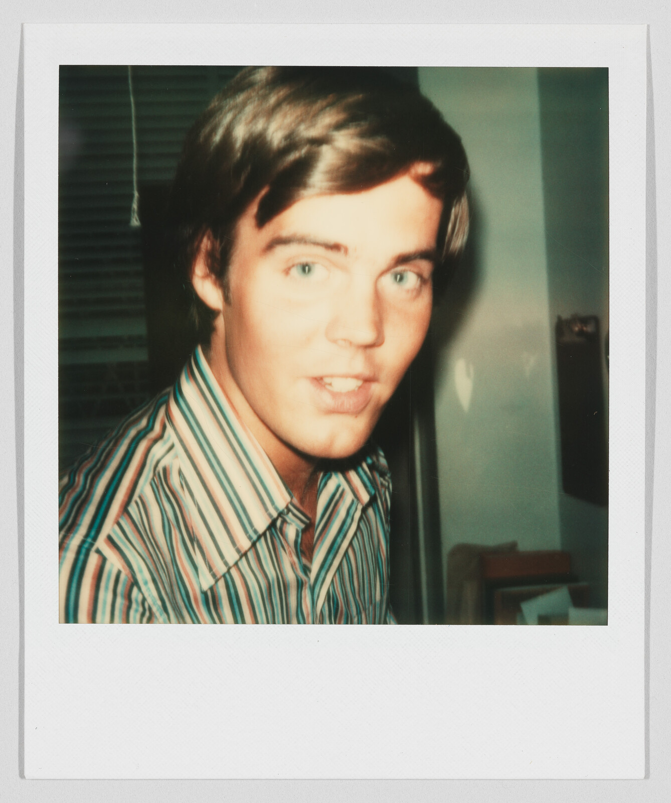 Young man with light brown hair wearing a striped shirt, looking toward the camera indoors.
