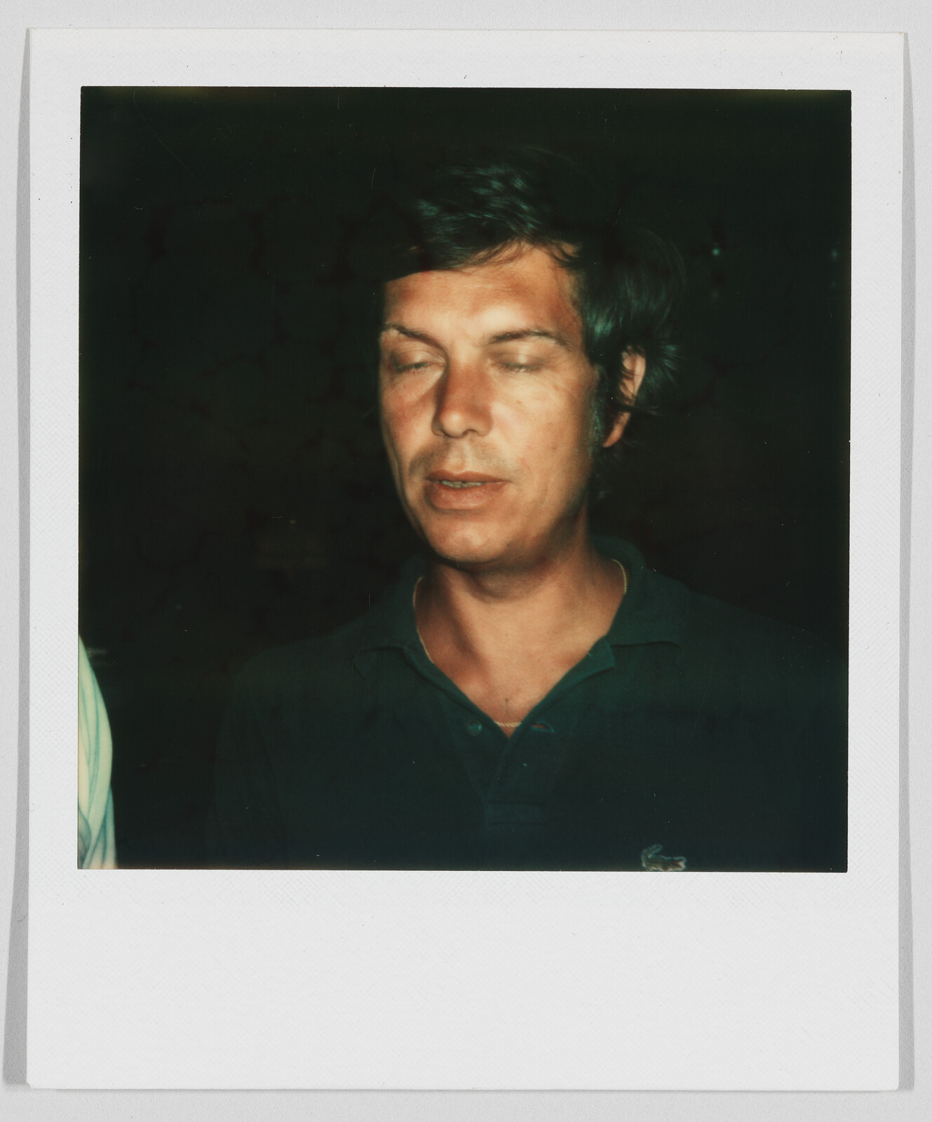 Man with short dark hair wearing a dark shirt, eyes closed, standing against a dark background.