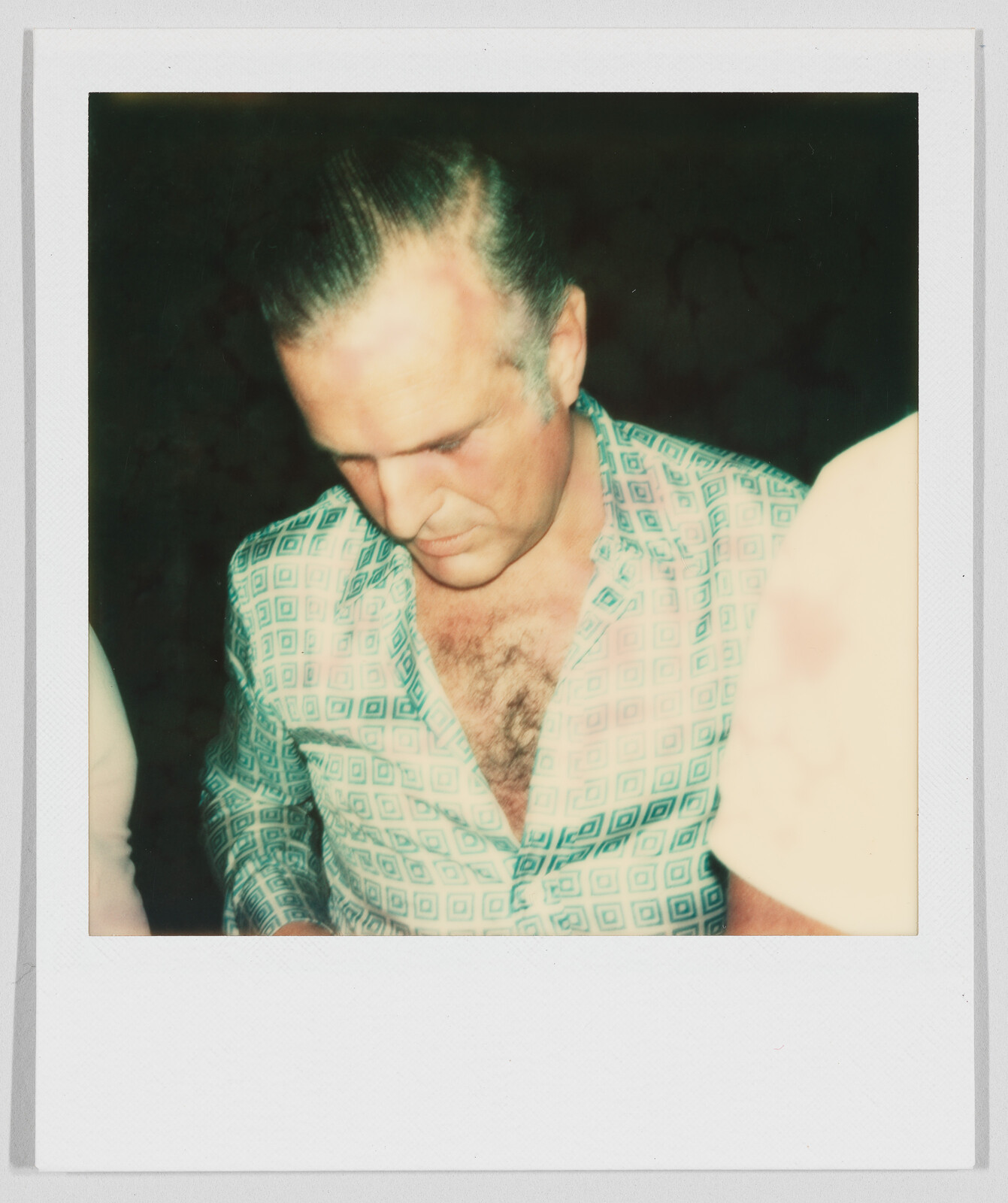 Man with slicked-back hair looks down, wearing a patterned shirt with an open collar, chest visible.