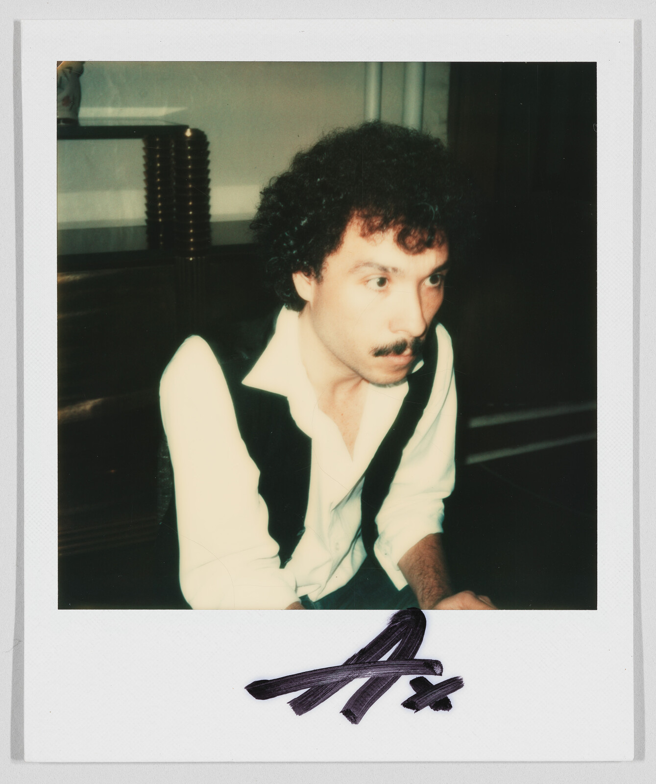 Man with curly hair and mustache wearing a white shirt and black vest, with “A+” written below.