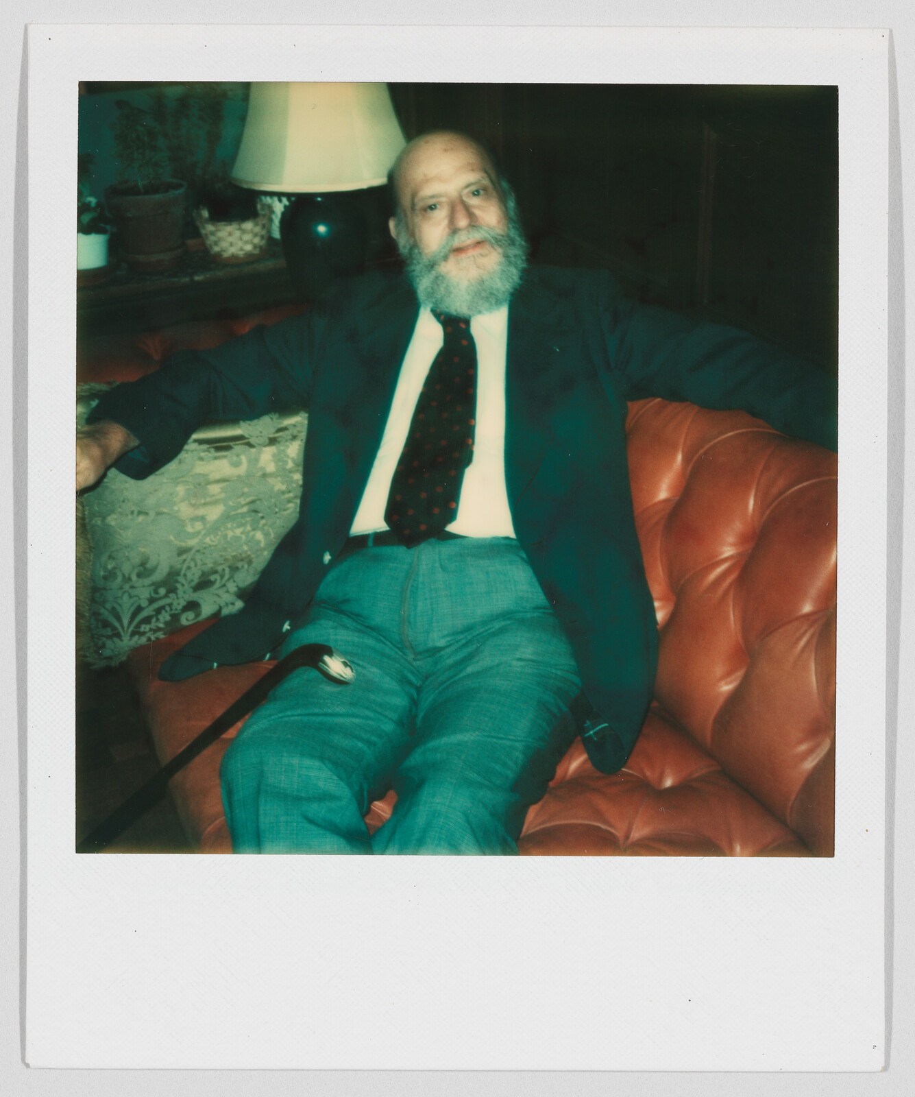 Older man with a white beard sits on a red couch, wearing a suit and holding a cane.