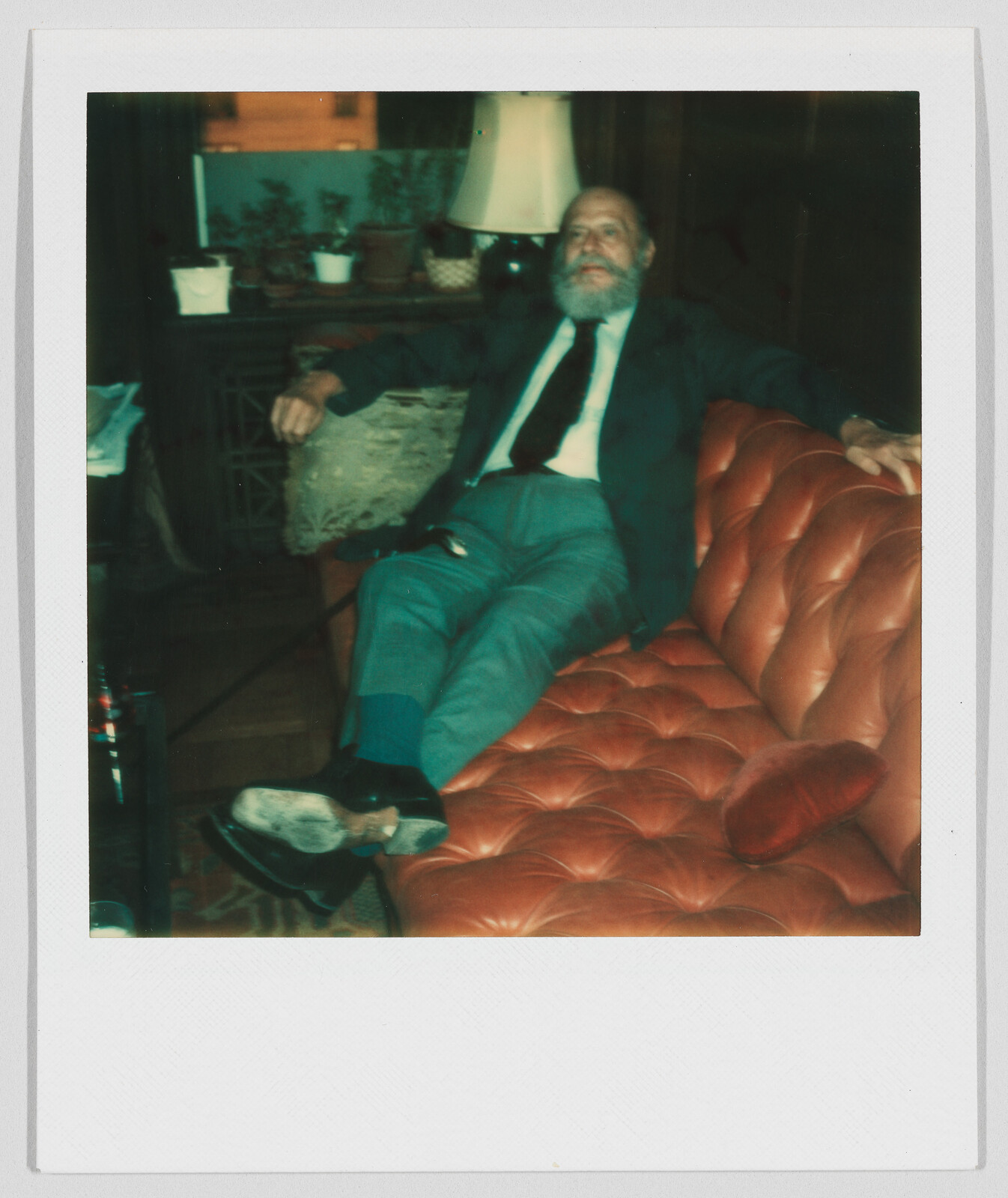 Older man with a beard sits relaxed on a red couch, wearing a suit and tie.