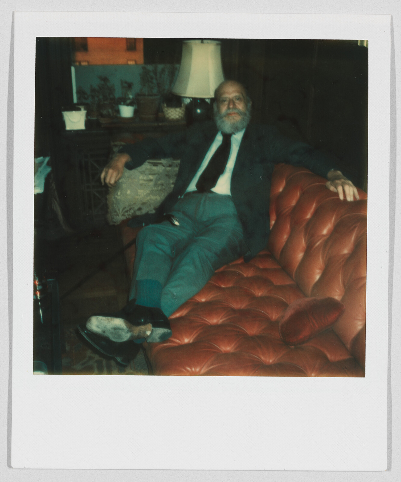 An older man with a beard sits relaxed on a red leather couch, wearing a suit and tie.