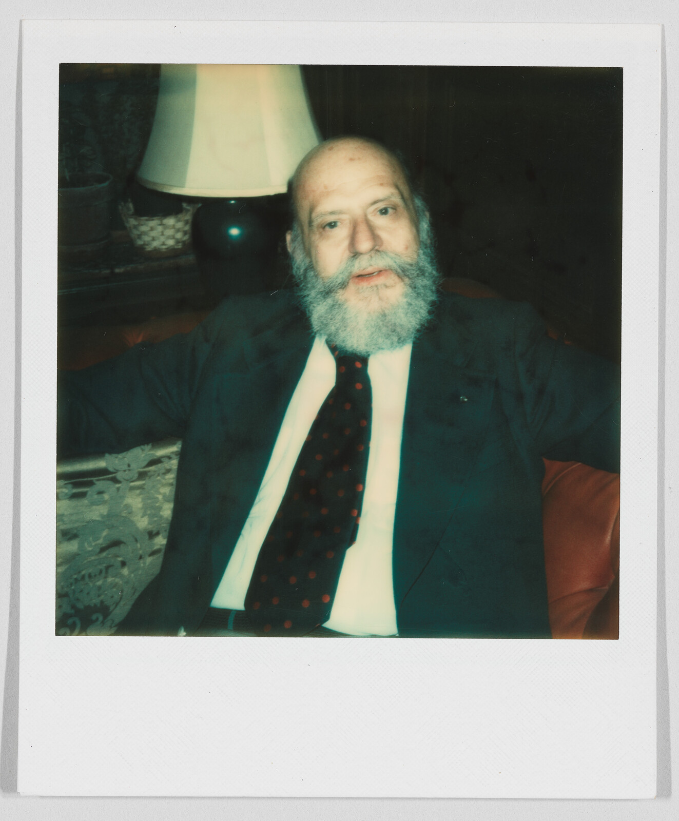 Older man with a gray beard wearing a suit and tie sits on a couch with arms resting wide.