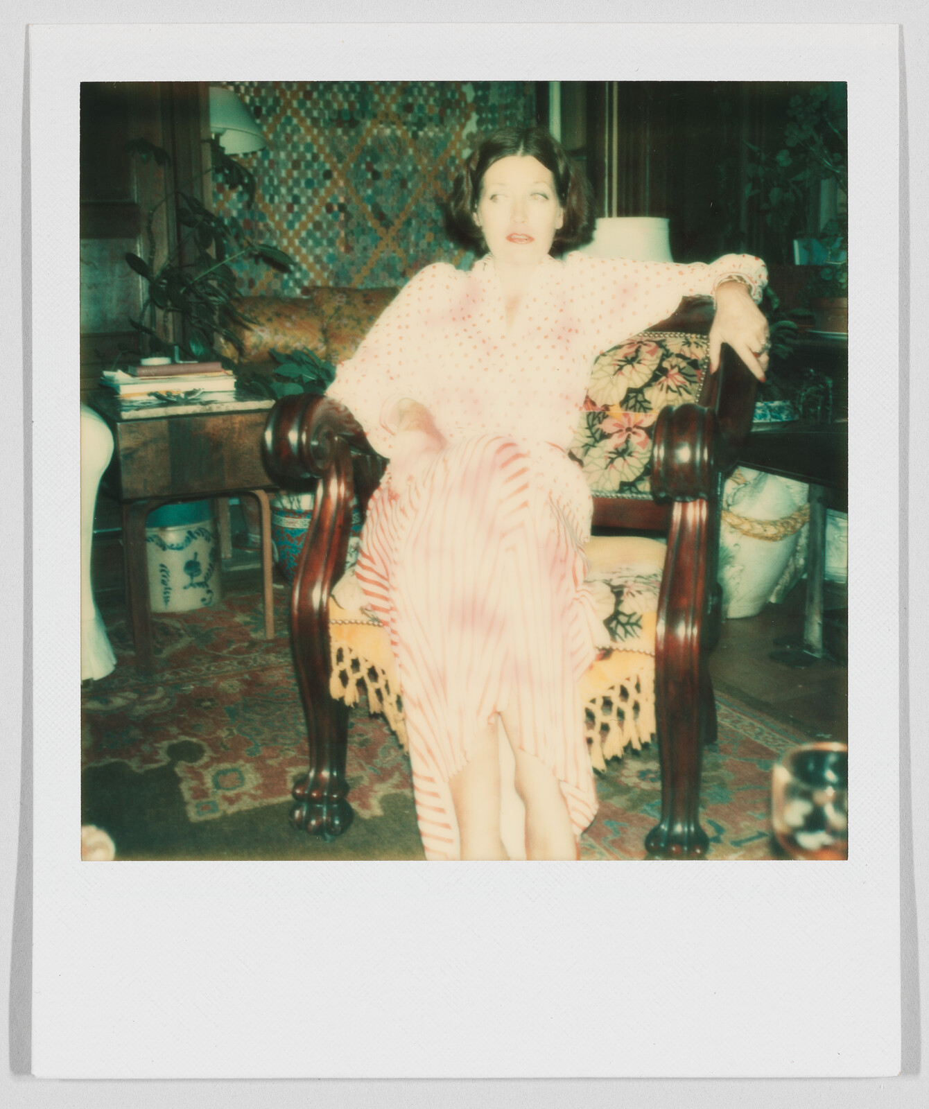 Woman in a long, light-colored dress sits on a wooden chair in a cozy, decorated living room.