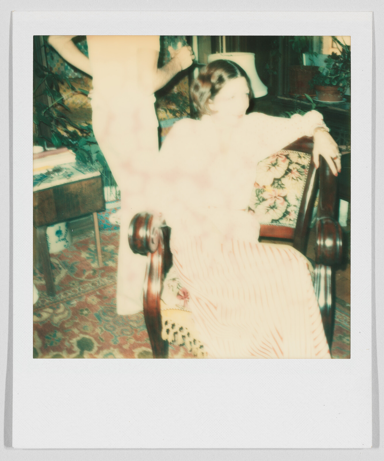 A person sits on a patterned chair while another stands nearby in a softly lit, vintage-style room.