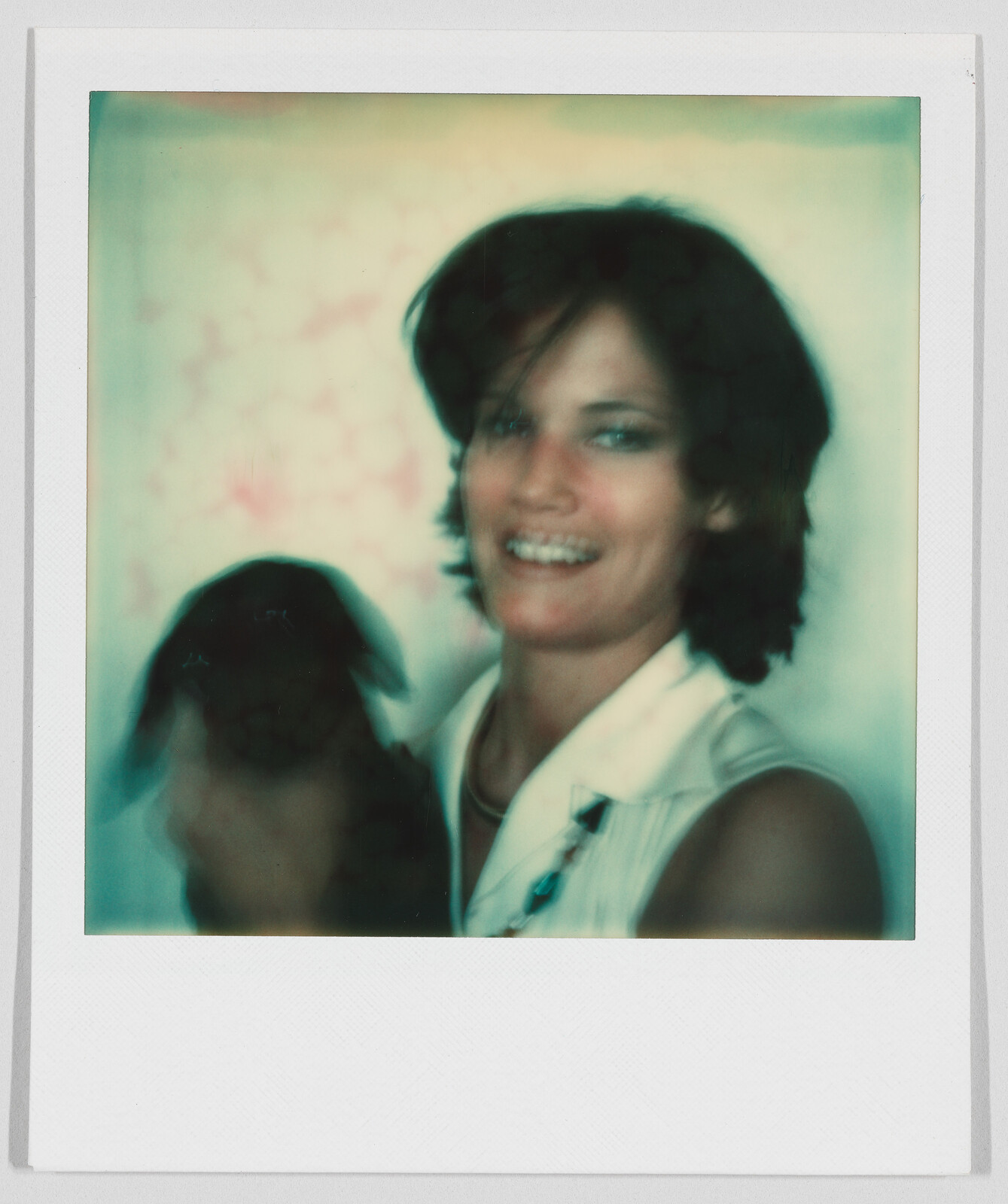 Smiling woman with short dark hair holds a small, blurry black animal, possibly a dog, indoors.