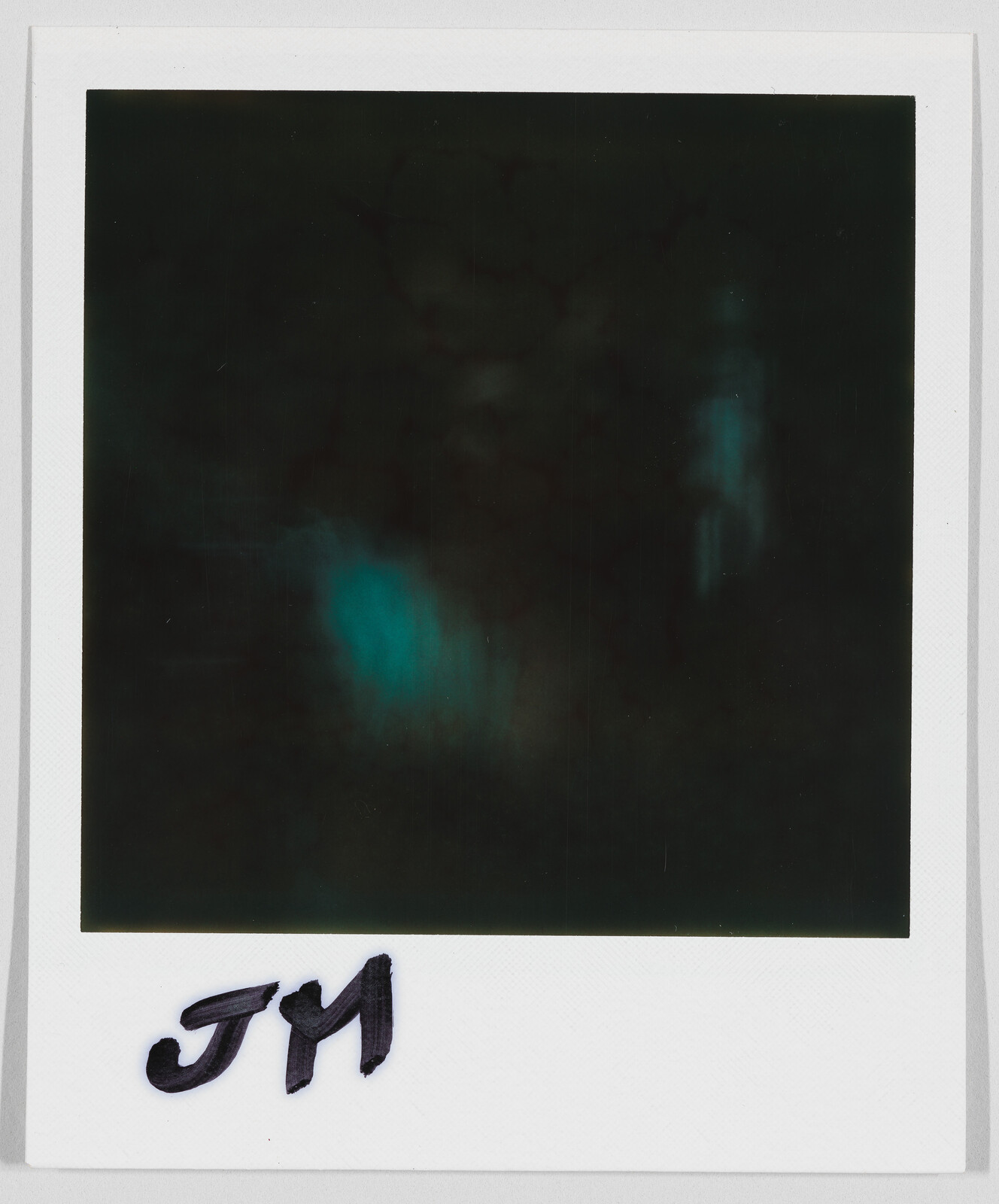 A dark, unclear Polaroid photo with the handwritten initials "JM" at the bottom border.