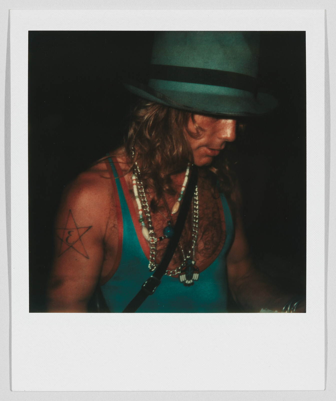Person wearing a blue tank top, beaded necklaces, and a hat, with a star tattoo on their shoulder.