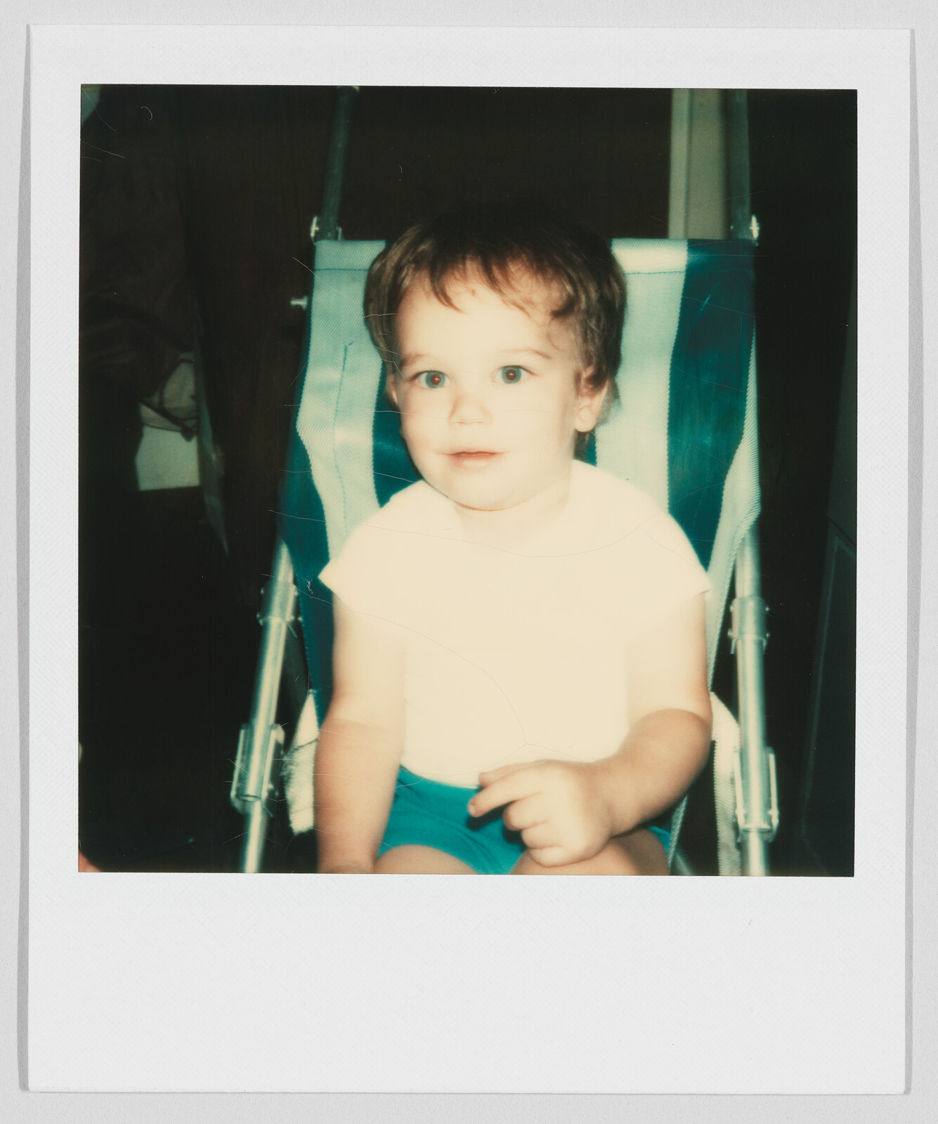 Young child with short hair sits in a stroller, wearing a white shirt and blue shorts, looking forward.
