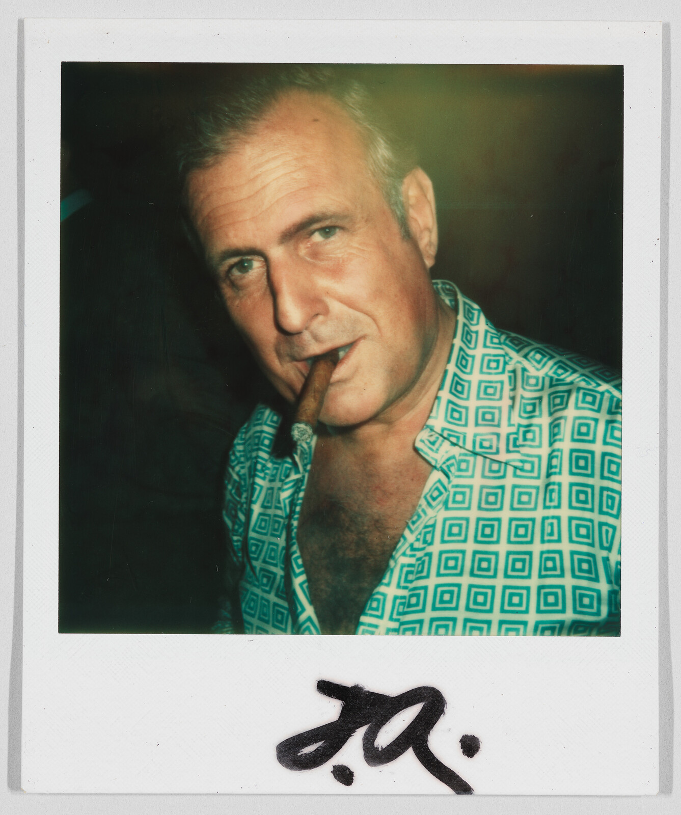 Man with short hair wearing a patterned shirt, holding a cigar in his mouth, with a signature below.