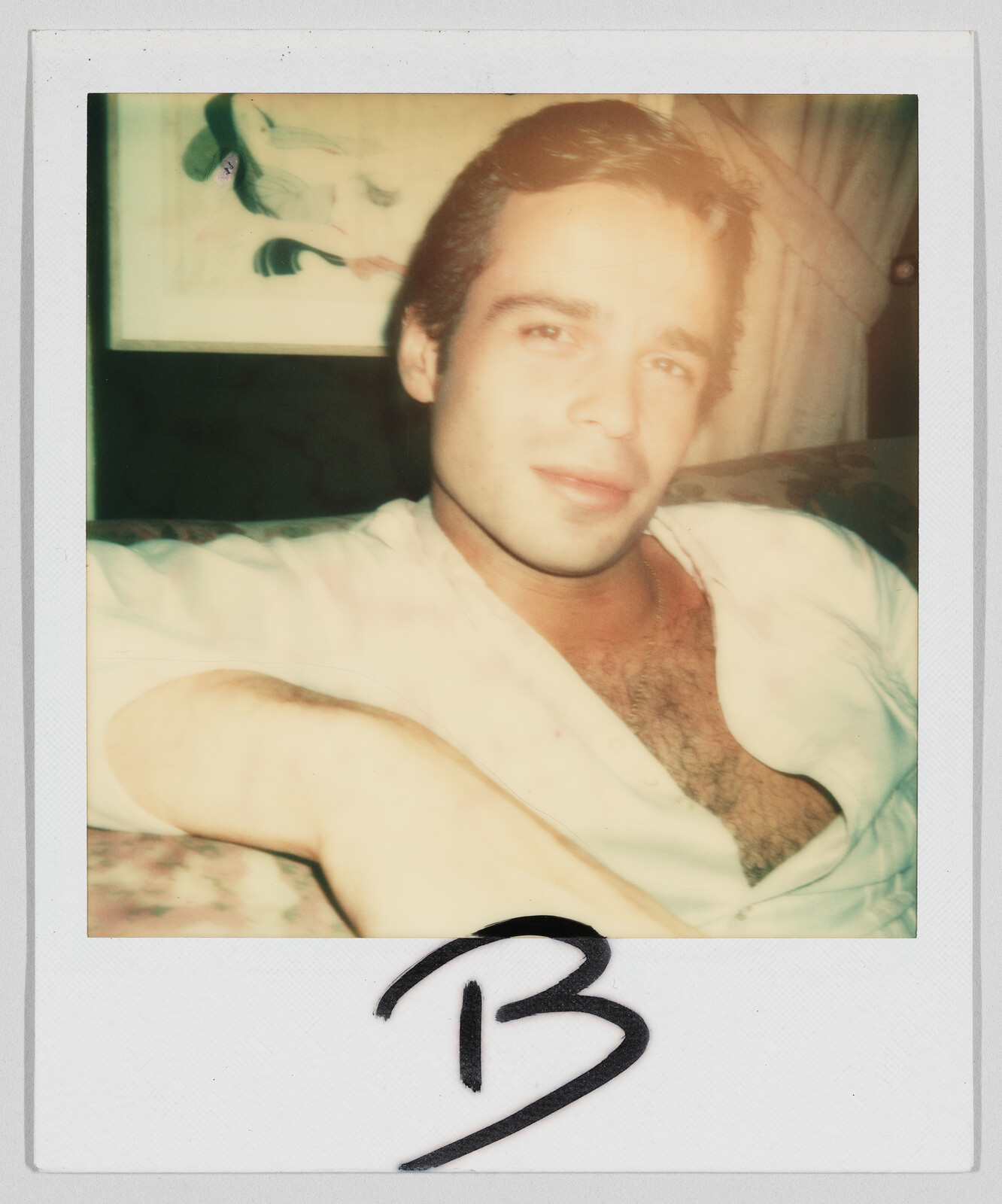 A man with short hair and an open shirt sits on a couch. The letter "B" is written below.