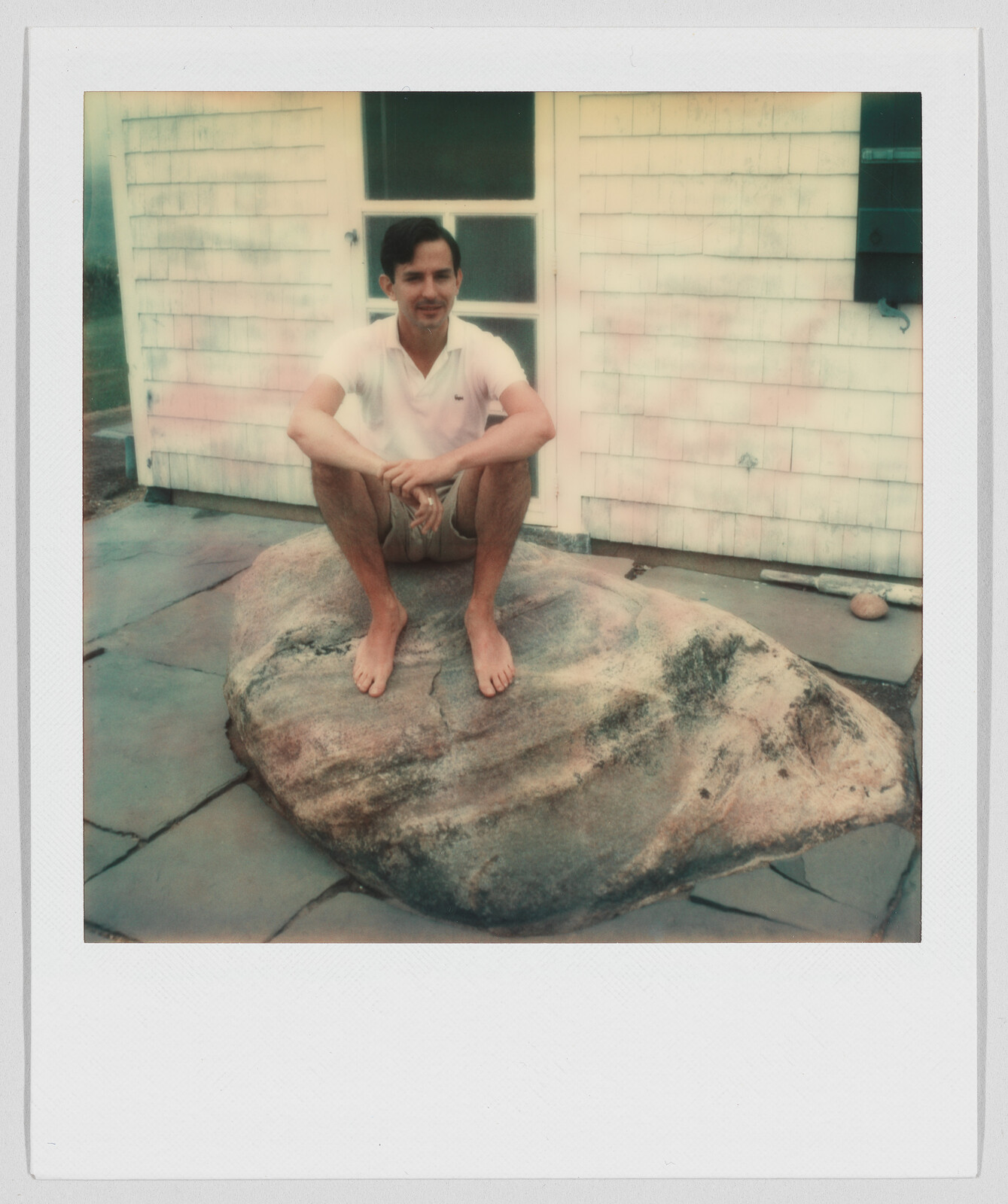 A man in a white shirt sits barefoot on a large rock in front of a house.