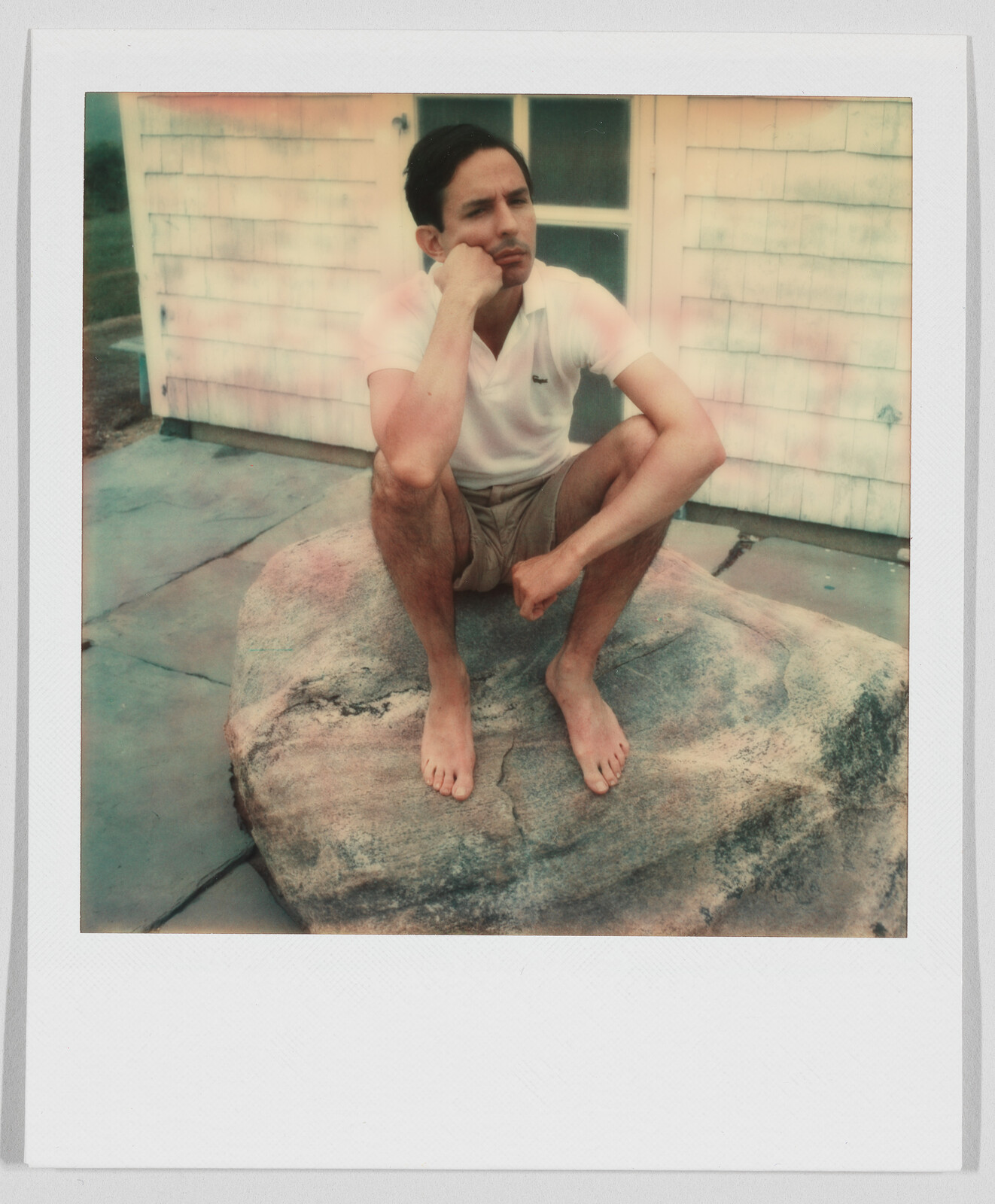 A barefoot man in a white shirt and shorts sits on a large rock, resting his chin on his hand.