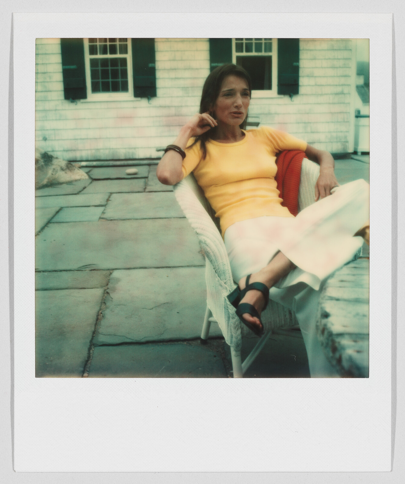 A woman in a yellow shirt and white pants sits relaxed on a patio chair outdoors.