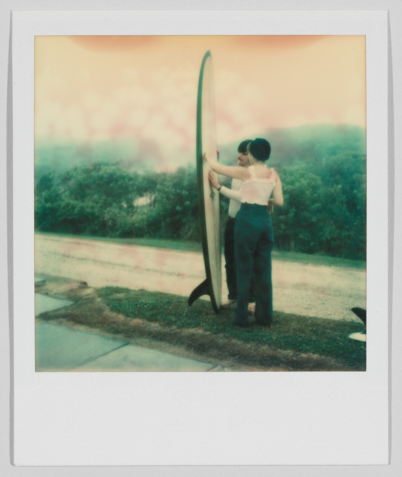 Two people stand outside, holding a tall surfboard together, with trees and a road in the background.