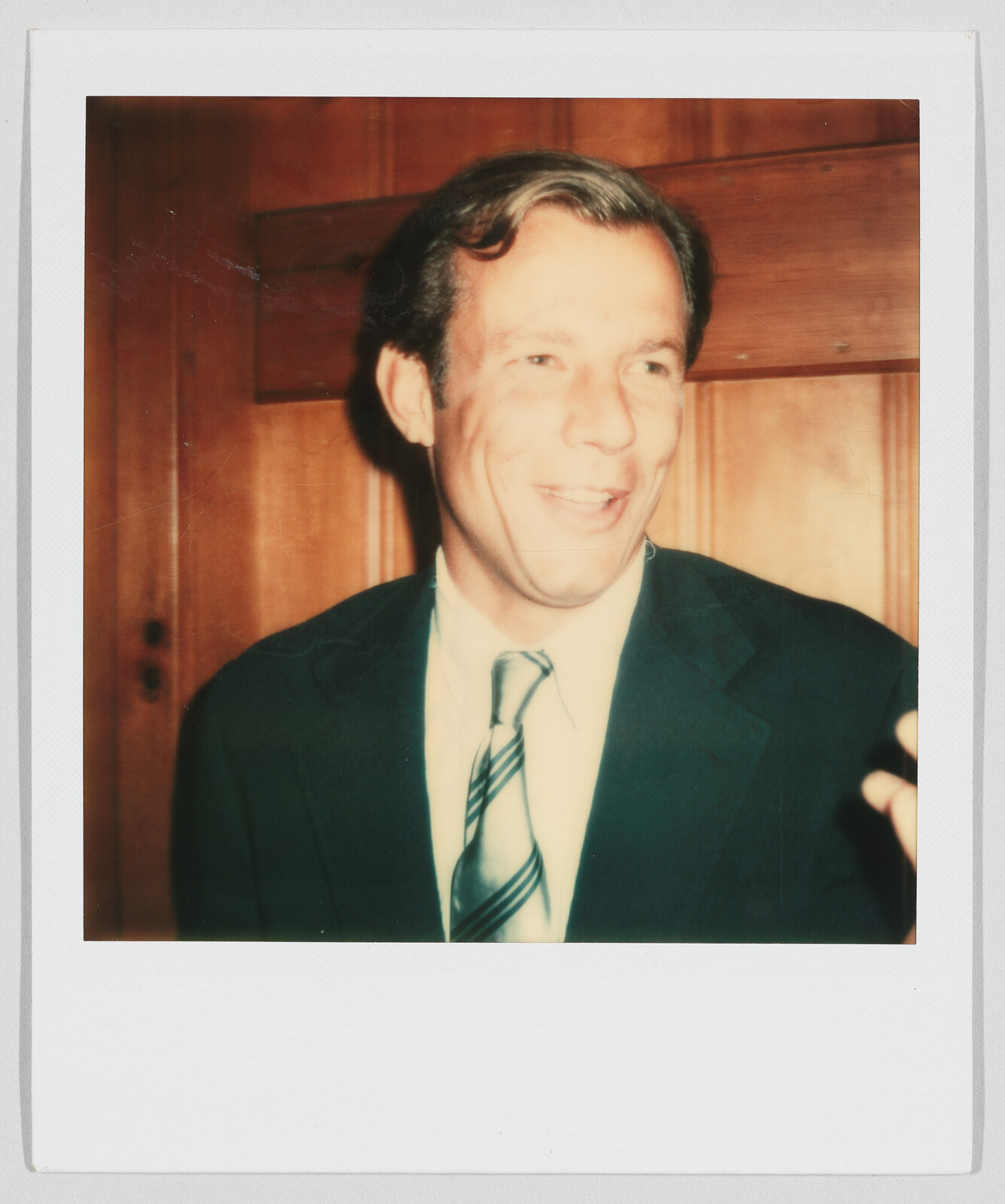 Man in a suit and striped tie smiling, standing in front of a wooden wall indoors.