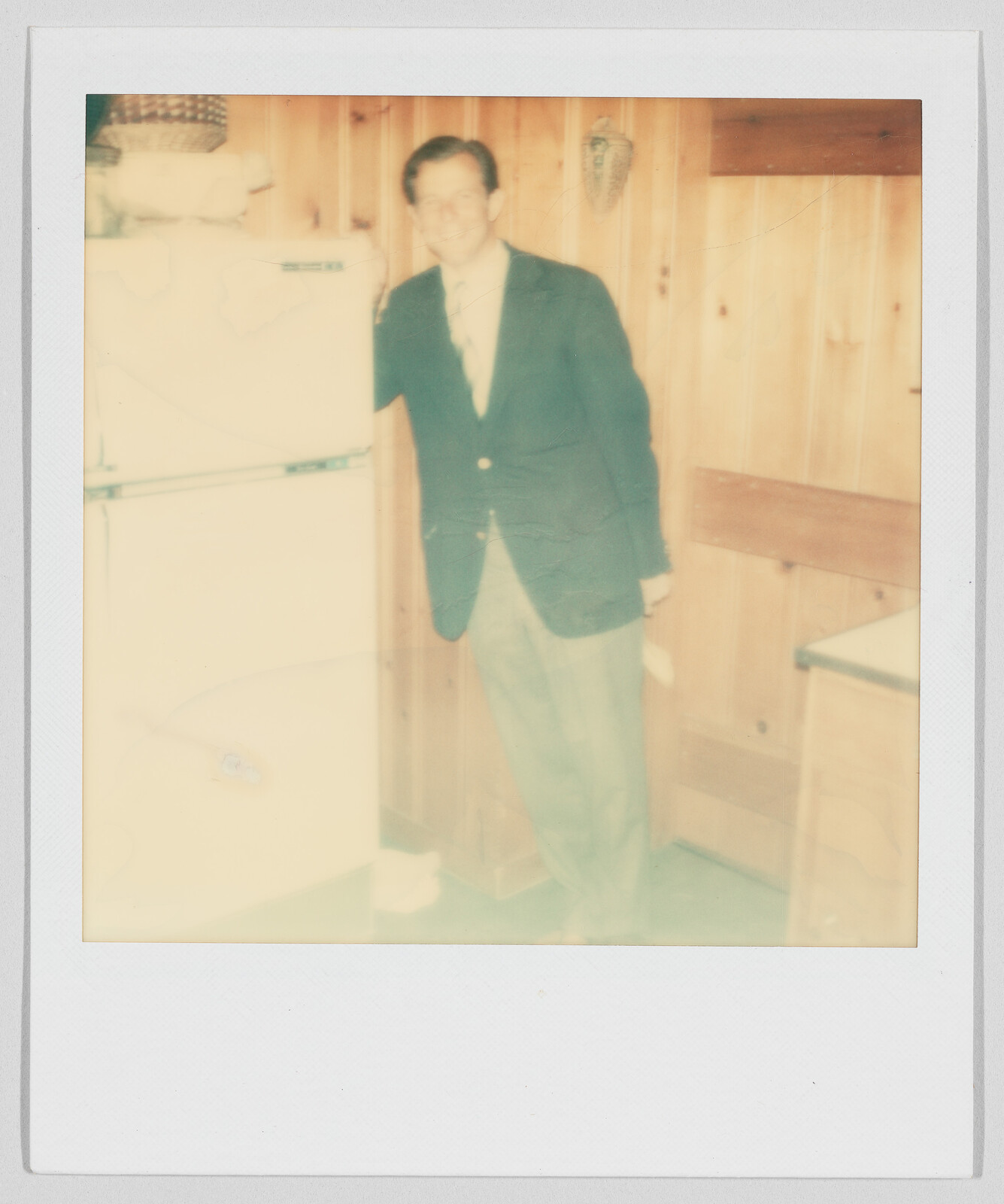A man in a suit stands next to a refrigerator in a wood-paneled kitchen, smiling at the camera.