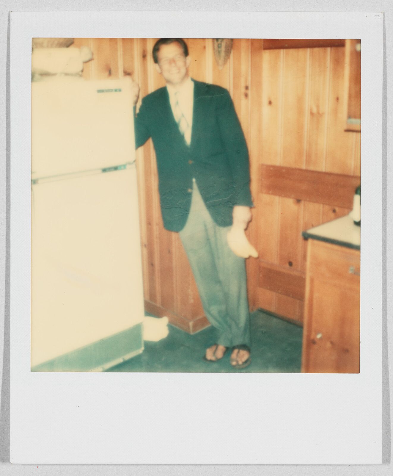 Man in a suit and sandals stands by a refrigerator in a wood-paneled kitchen, holding a banana.