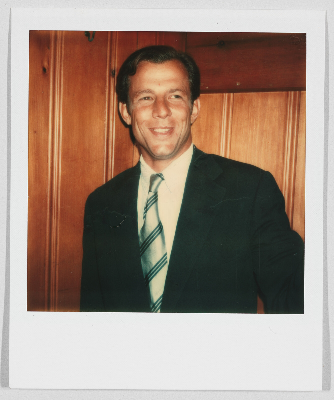 Man in a suit and striped tie smiling, standing in front of a wooden wall.