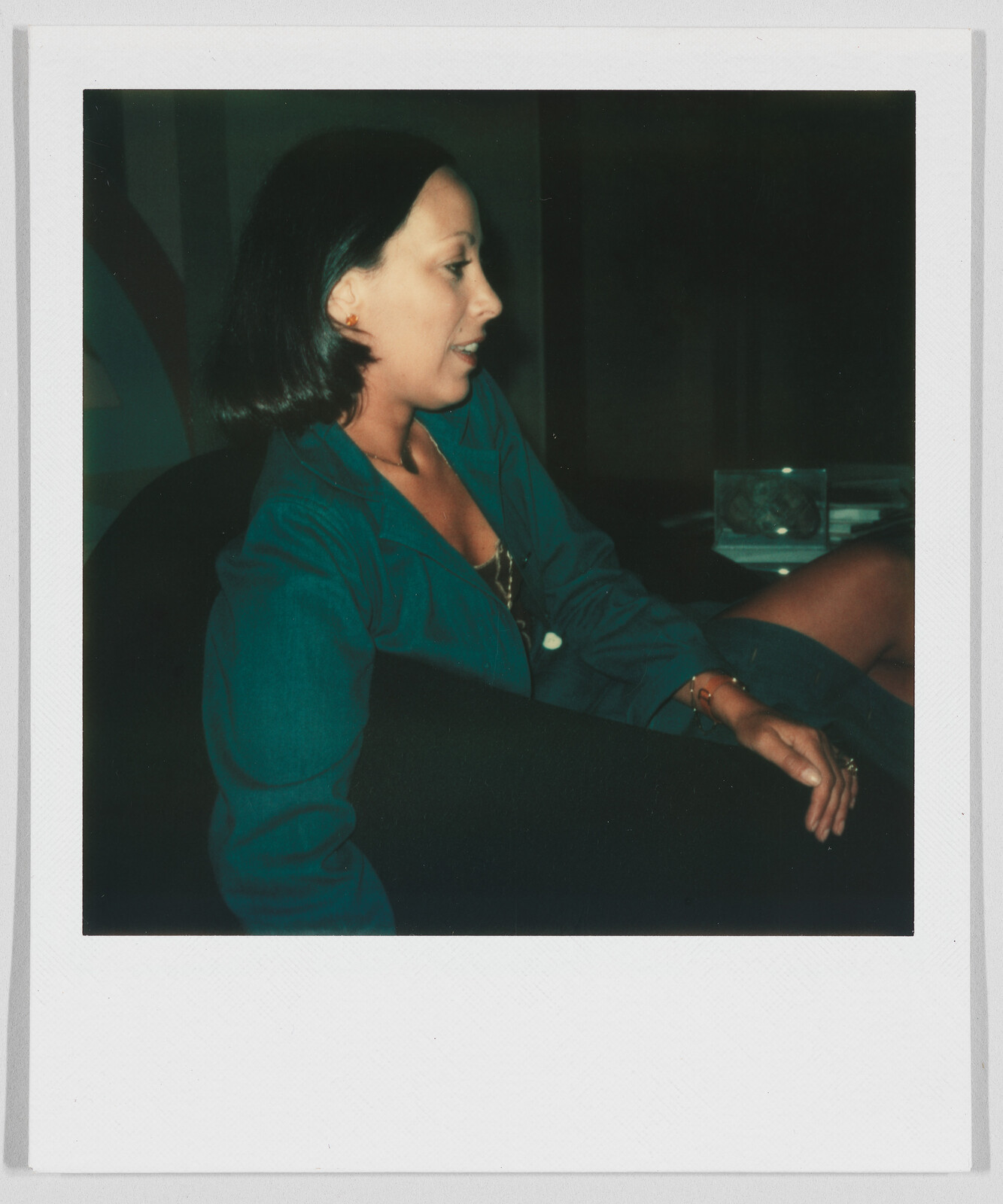 A woman with short dark hair sits sideways on a chair, wearing a teal jacket and looking to the left.