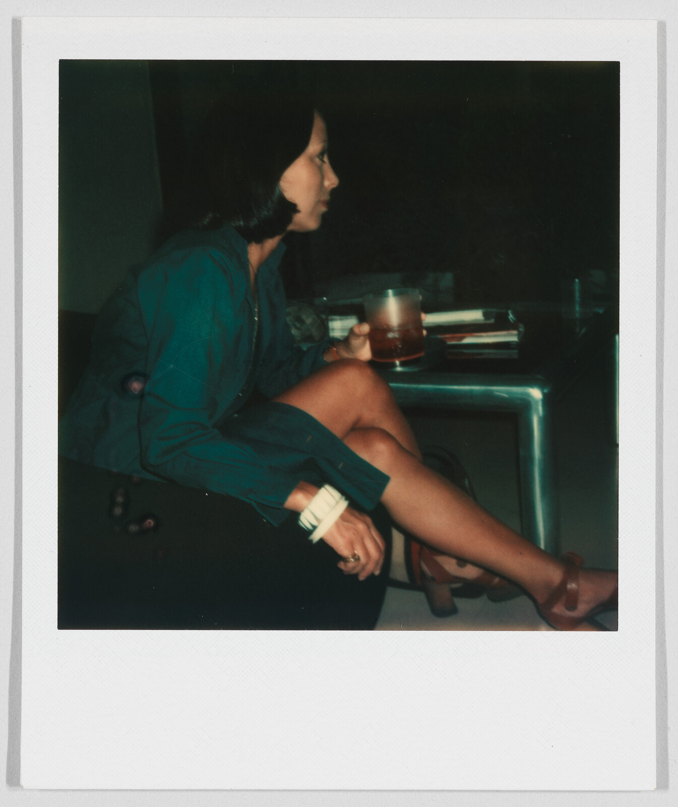 A woman sits on a couch holding a drink, looking to the side with her legs crossed.