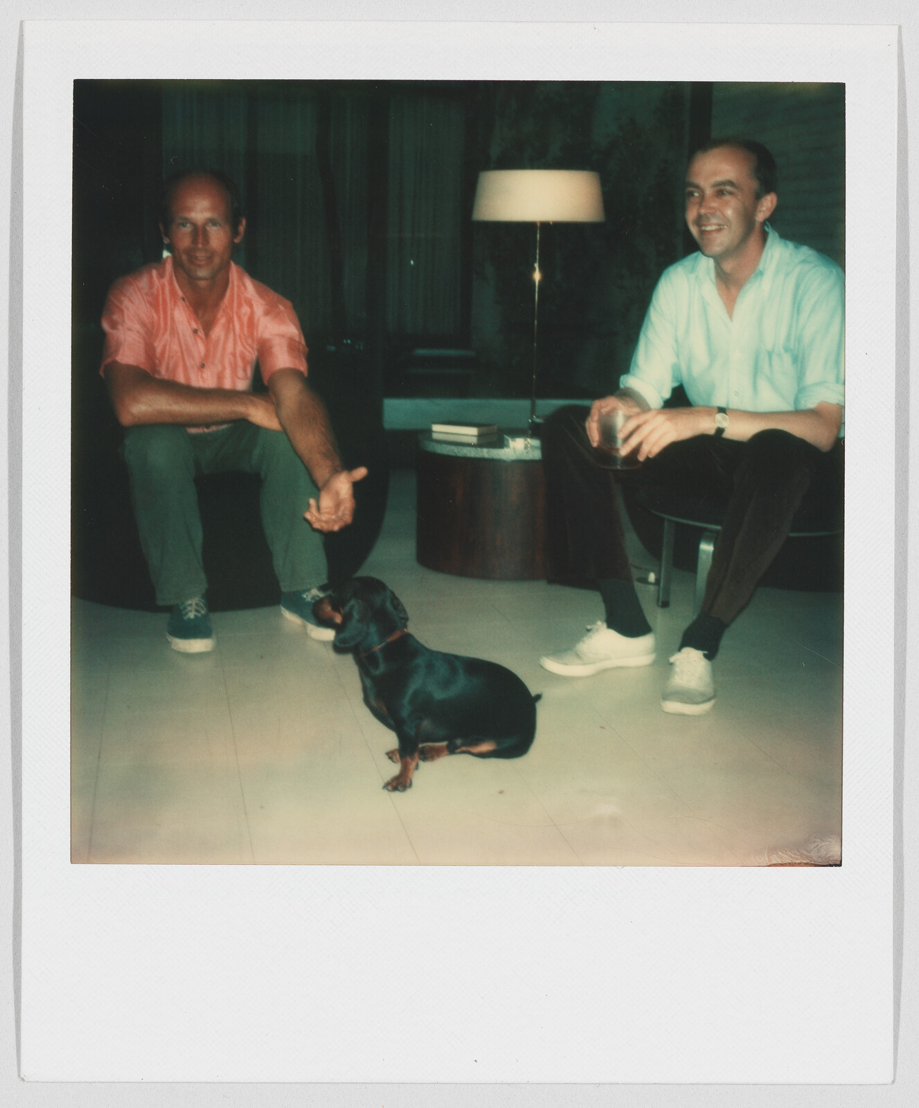 Two men sit indoors, smiling and looking at a small black dog sitting on the floor between them.