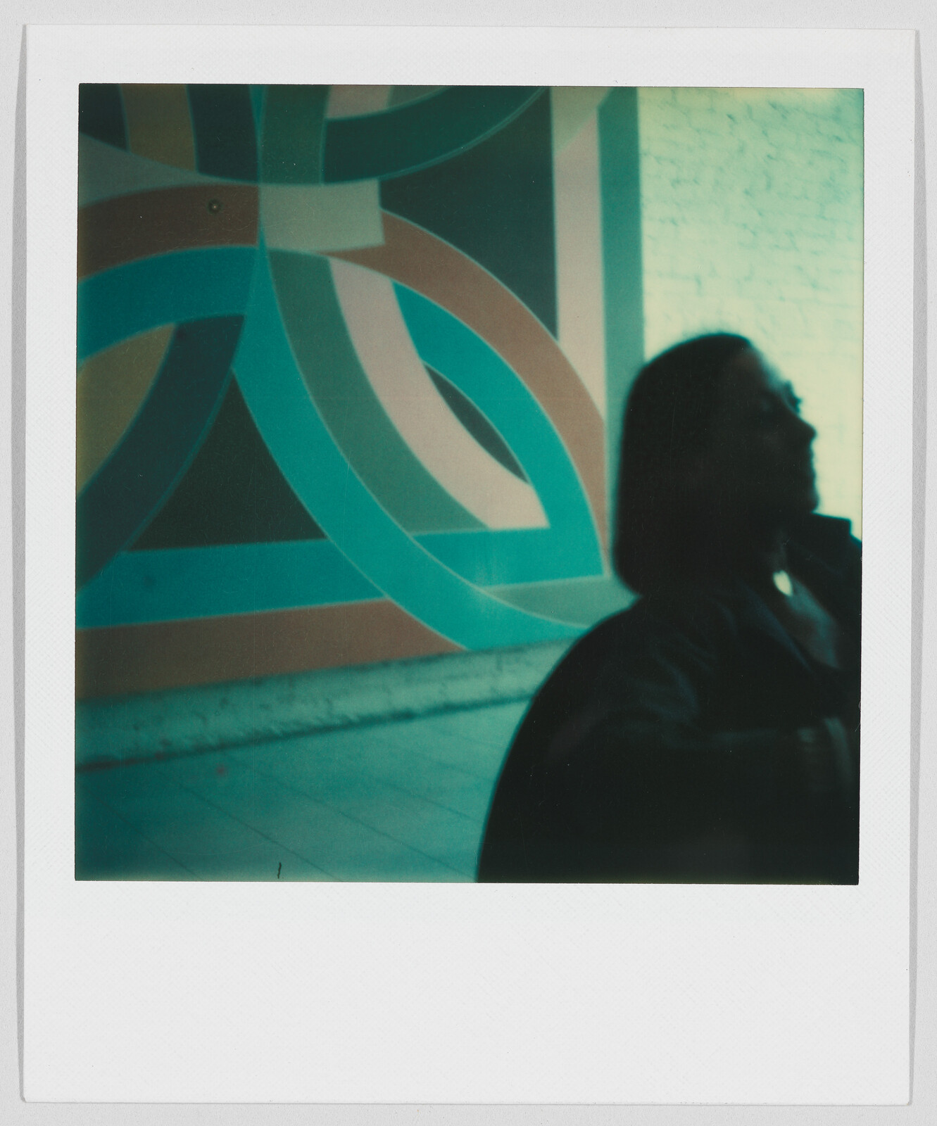 A person with shoulder-length hair stands in front of a colorful geometric mural on a wall.