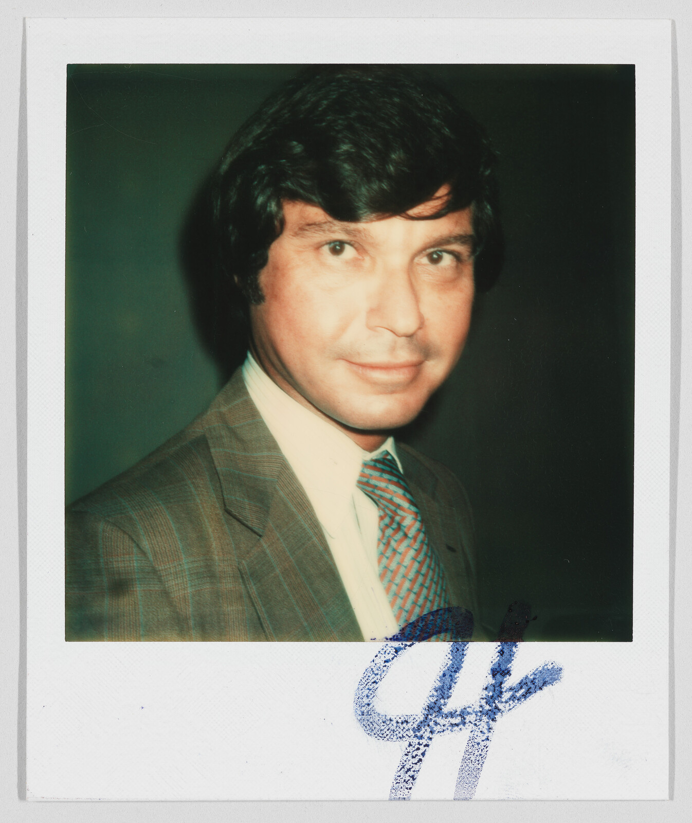 Man with dark hair wearing a plaid suit and patterned tie, with a large blue signature at the bottom.
