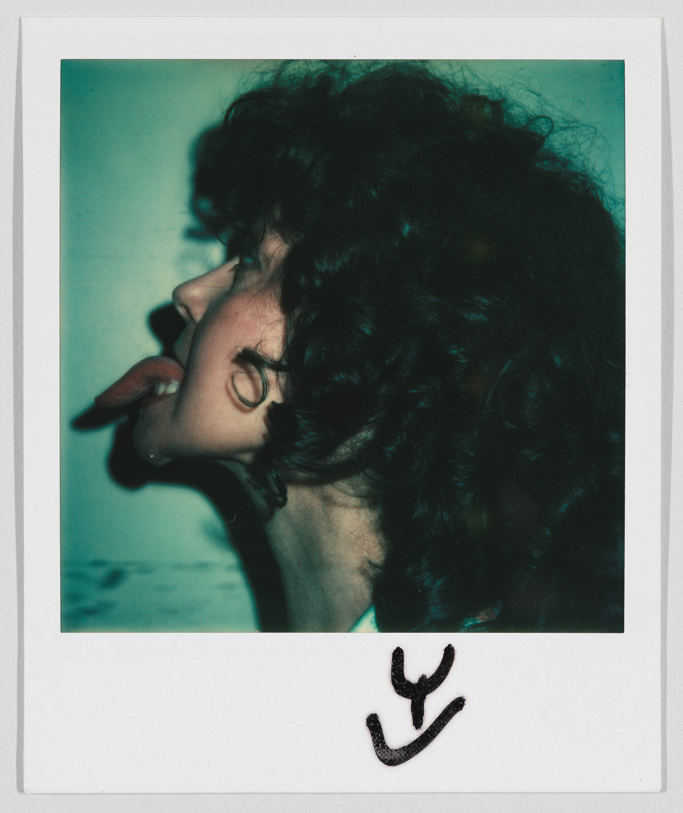 Person with curly hair sticking out their tongue, seen from the side. Black marker scribble at the bottom.