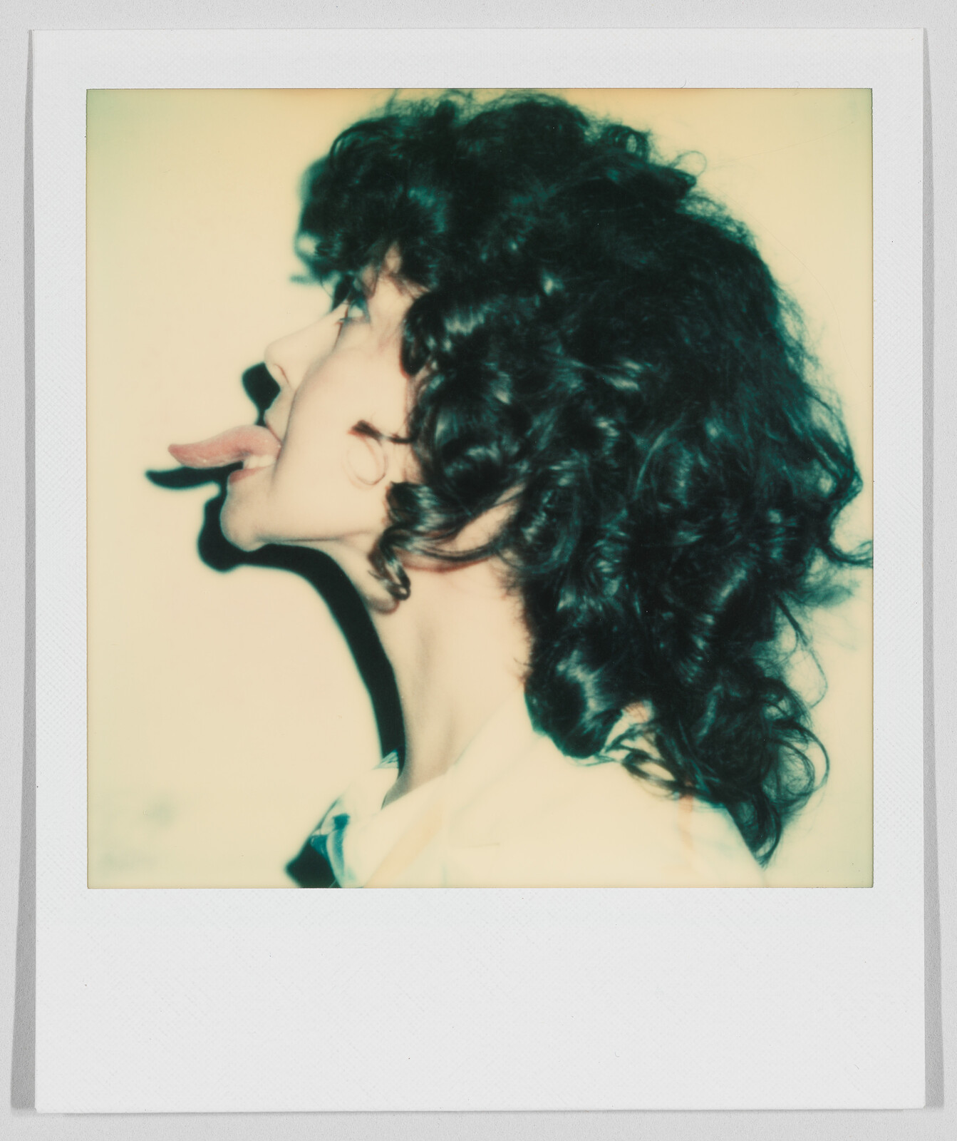 Person with curly hair in profile view, sticking out their tongue, wearing a light-colored shirt.