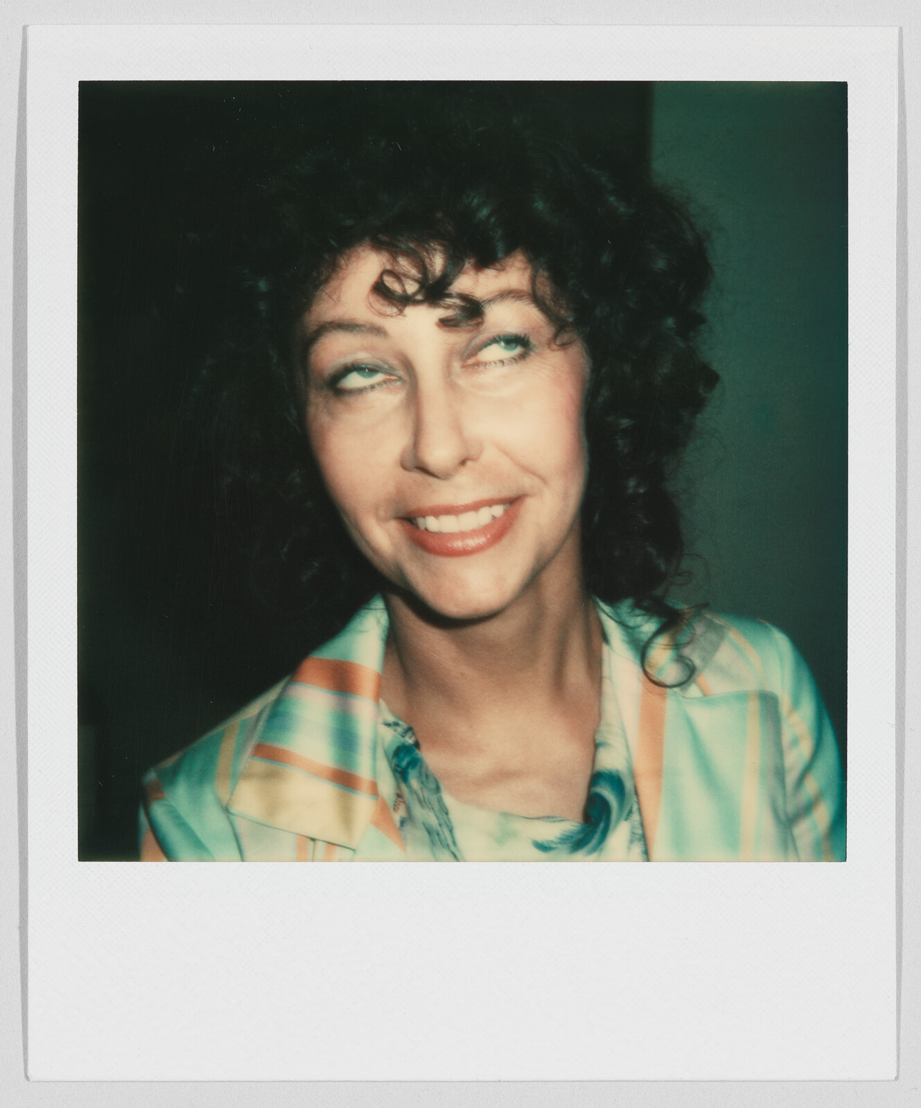 Woman with curly hair smiling and looking upward, wearing a colorful striped jacket over a patterned shirt.
