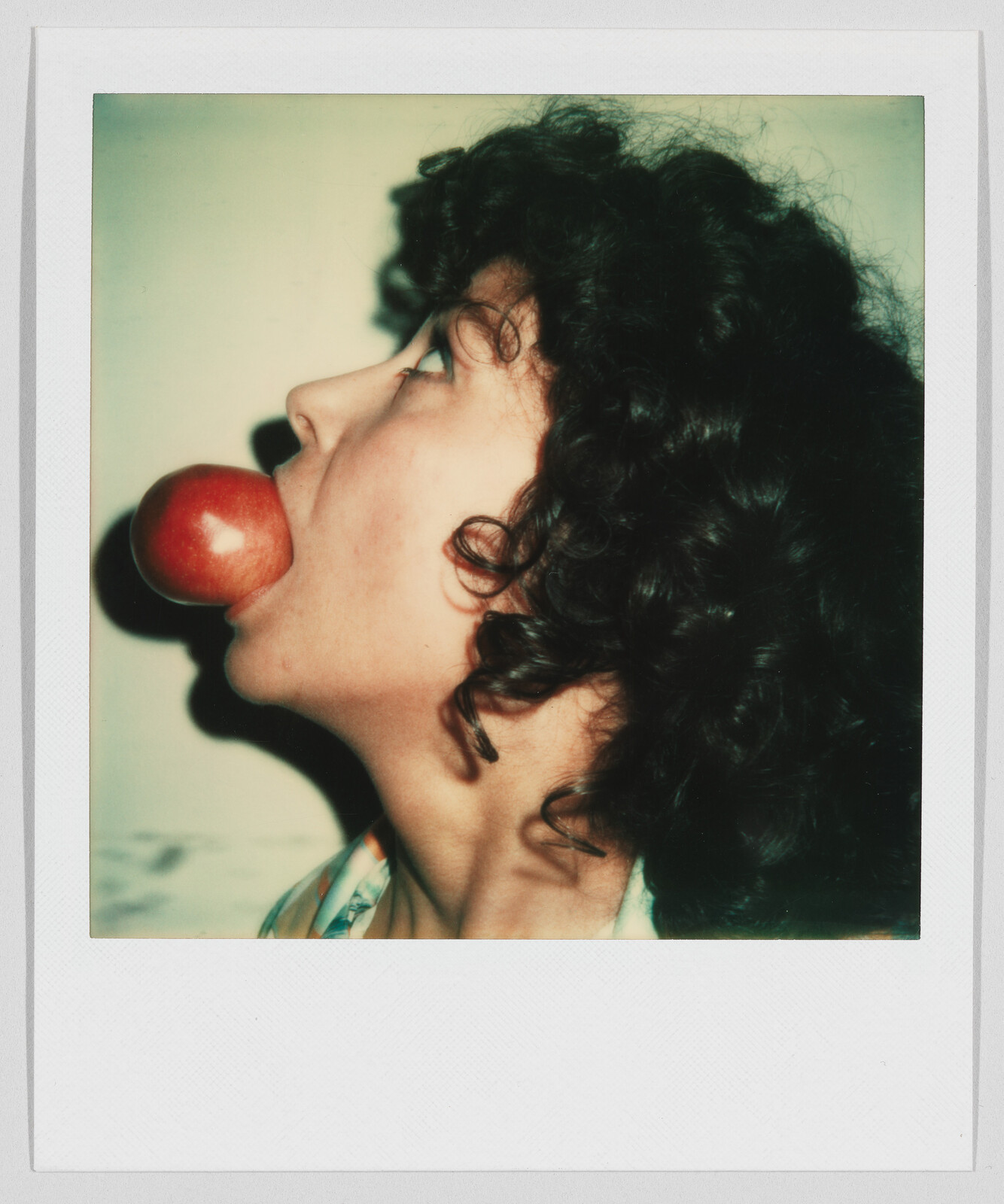 Person with curly hair holding a red apple in their mouth, head tilted back and mouth open wide.